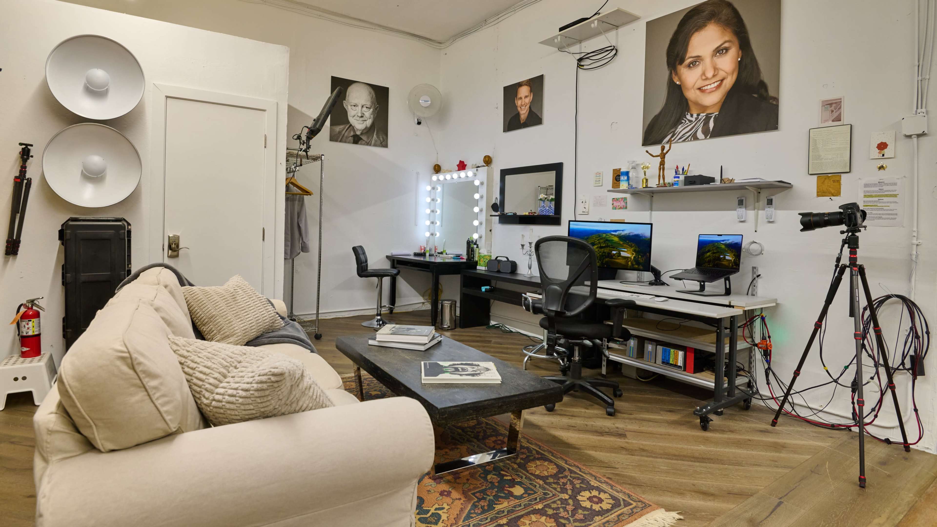 A studio workspace with two computer desks, a small sofa, a makeup mirror, and several photographs on the walls.