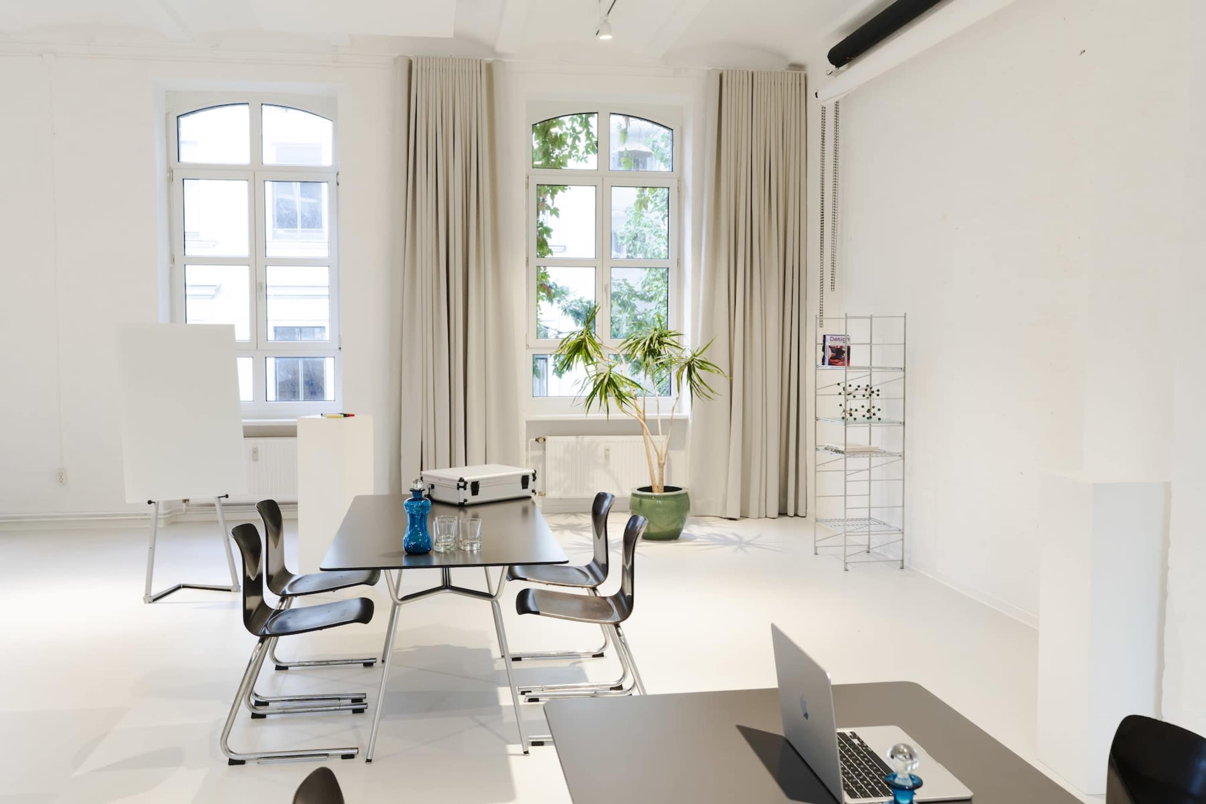 The image shows a modern, bright room with large windows, a potted plant, a folding table with chairs, and a workspace equipped with a laptop.