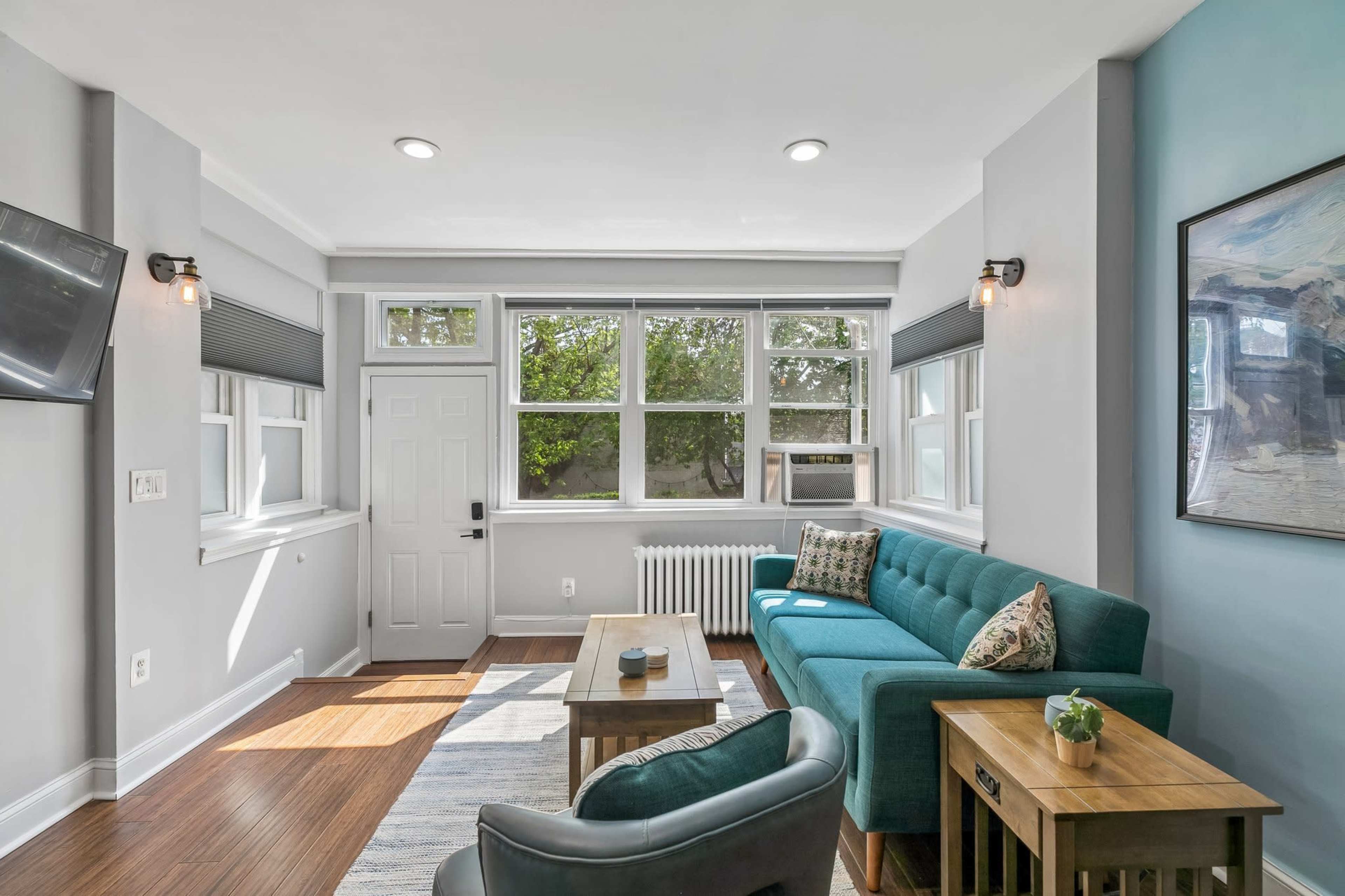 The image shows a well-lit living room featuring a teal sofa, a coffee table, and large windows with a view of greenery outside.