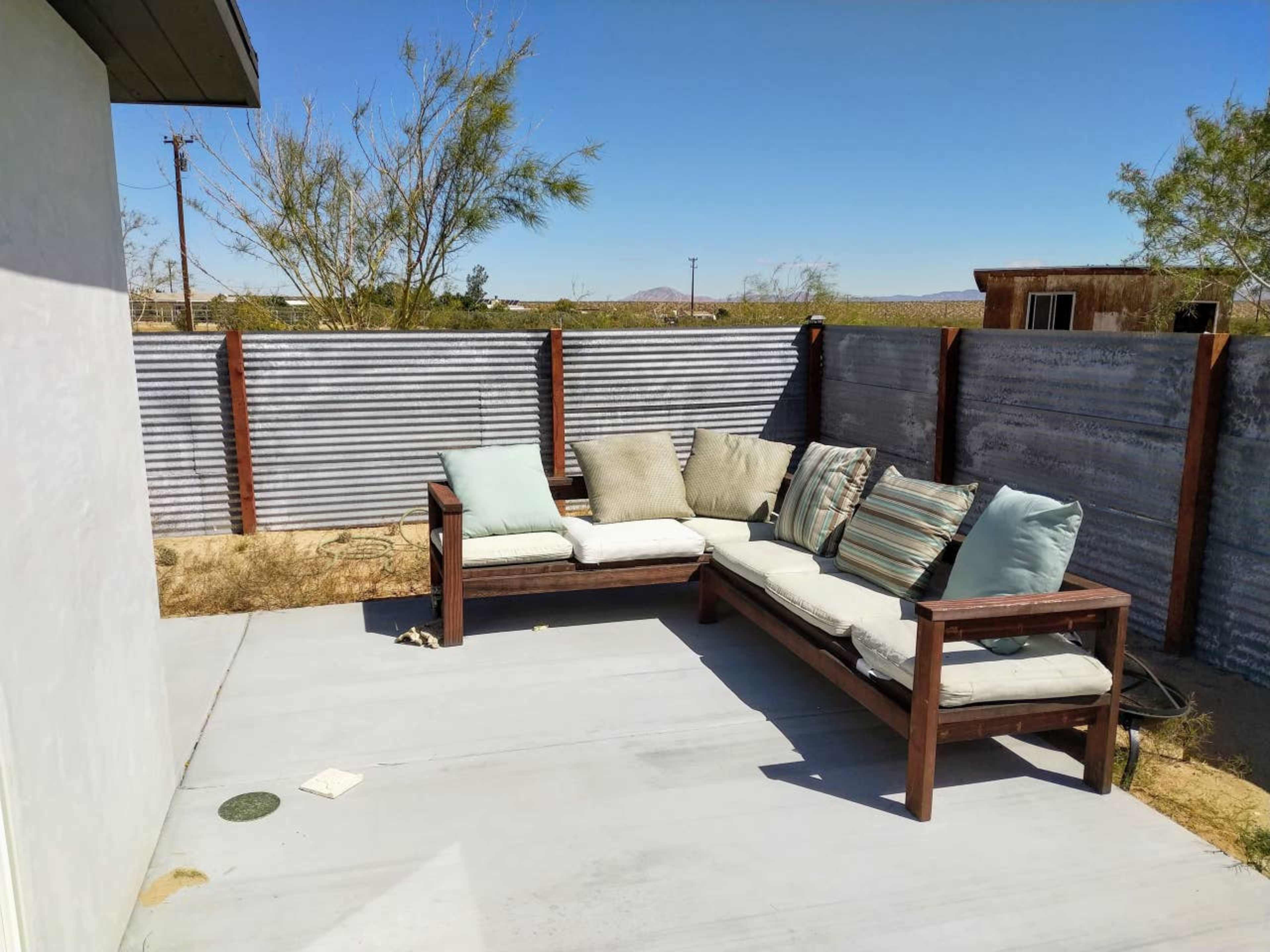 A wooden outdoor seating area with cushions, surrounded by a corrugated metal fence and sparse vegetation.
