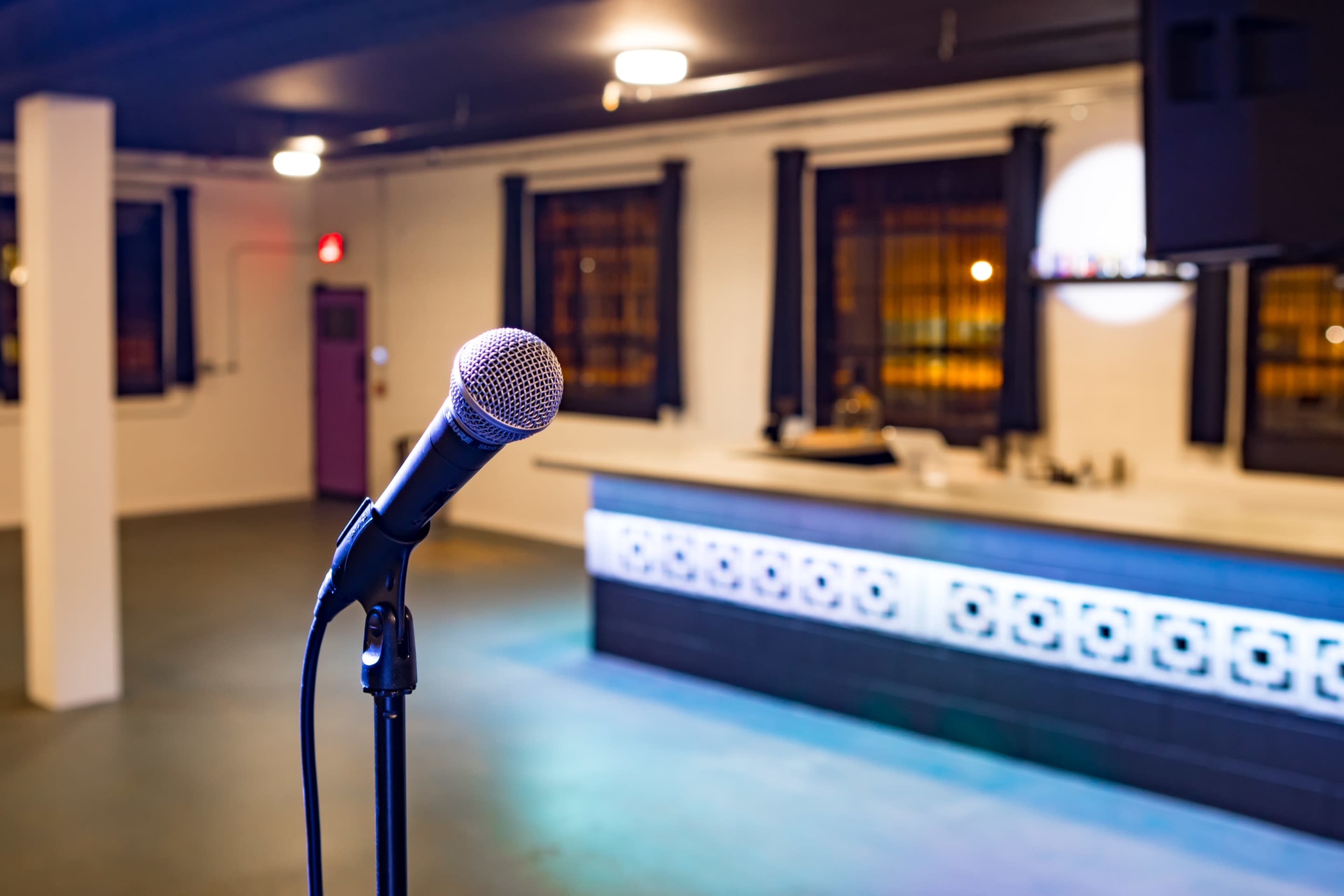 A microphone stands in the foreground, with a partially visible bar area and empty room in the background.