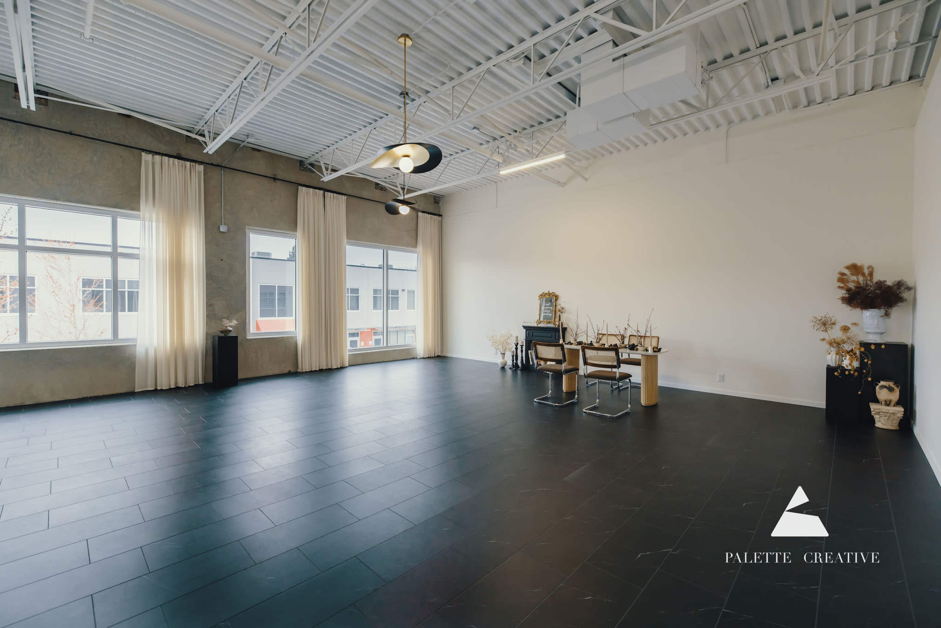 The image shows a spacious, well-lit room with large windows, featuring a modern table and chairs along with decorative elements.