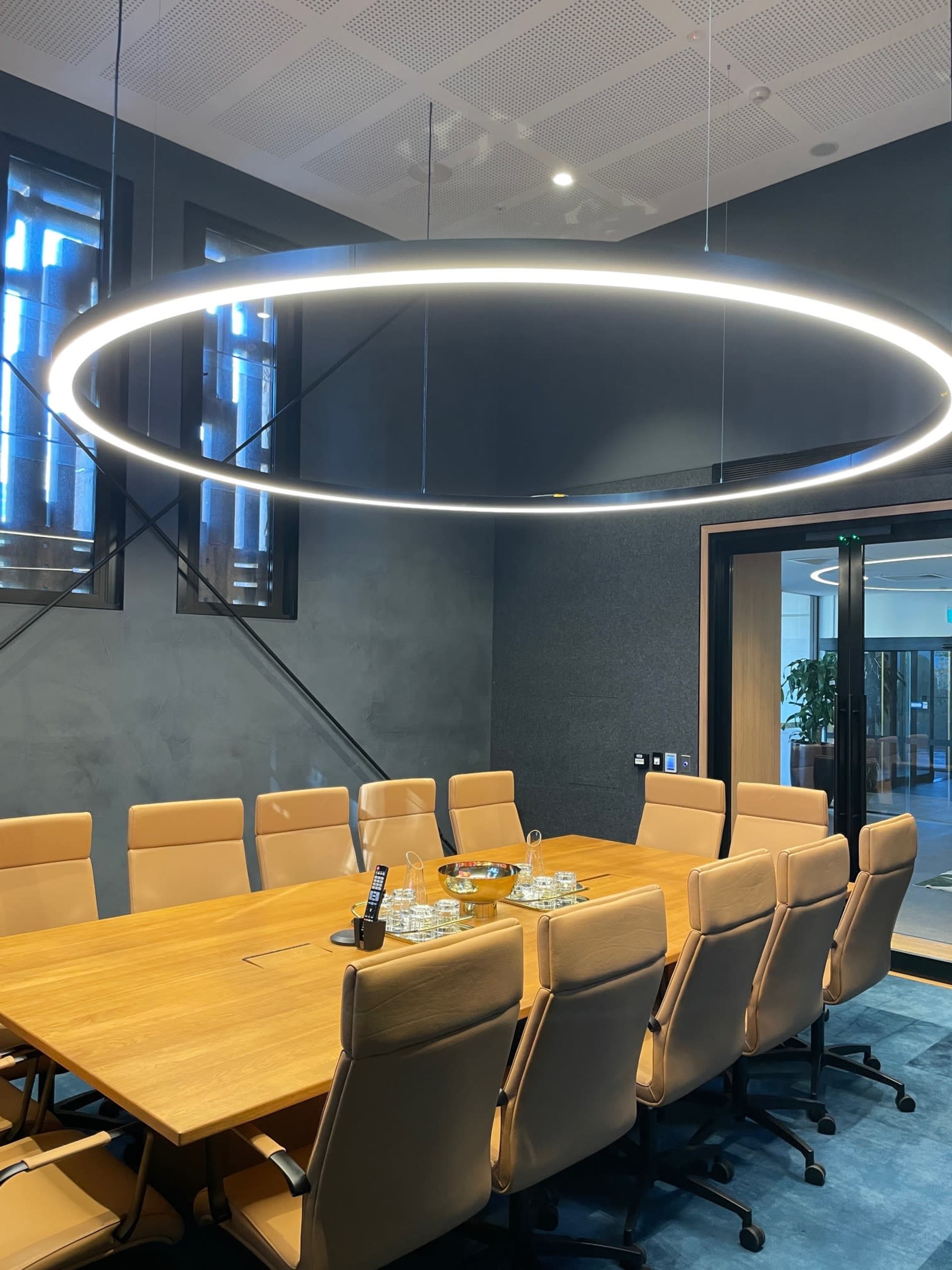 A modern conference room features a large wooden table surrounded by twelve leather chairs and an overhead ring light.
