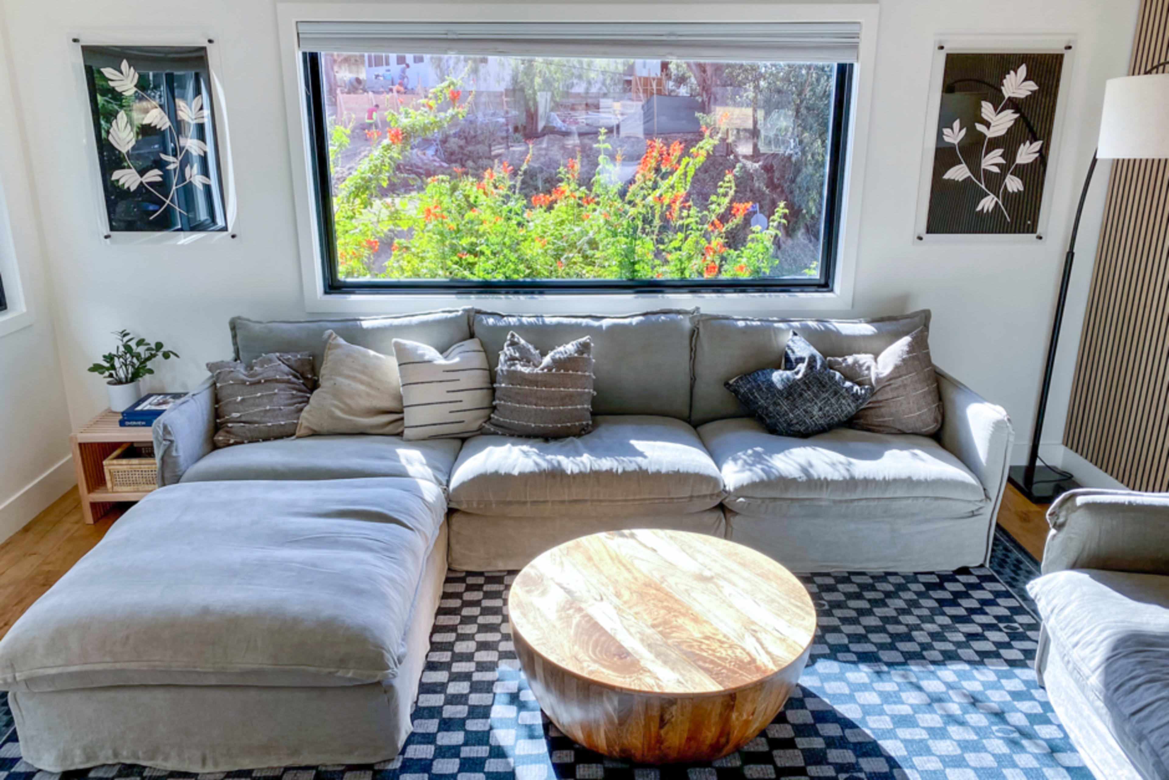 The image shows a cozy living room featuring a gray sectional sofa, a round wooden coffee table, and large windows revealing colorful greenery outside.