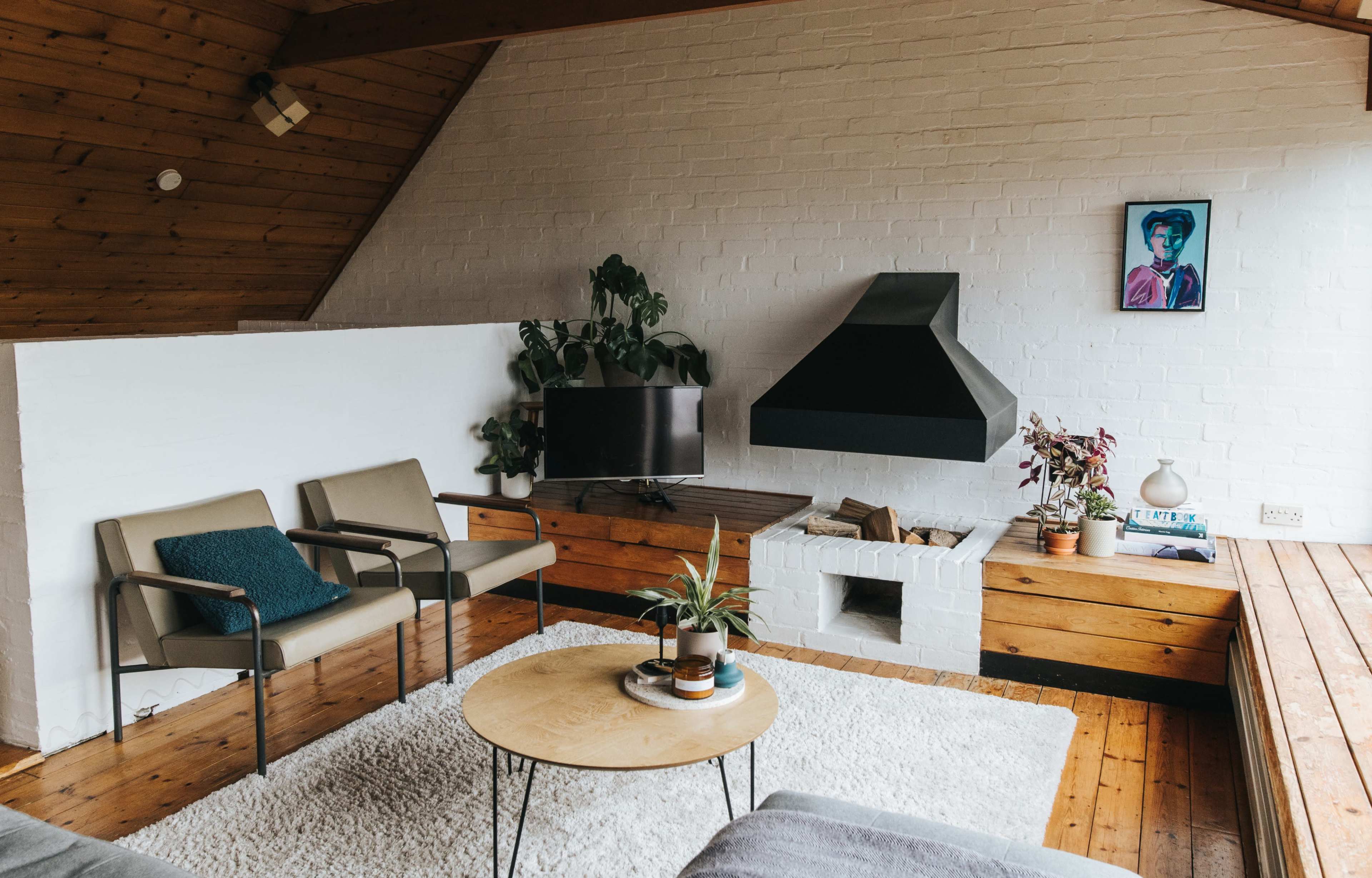 A cozy living room with two armchairs, a circular coffee table, a television, a fireplace, and plants, all set against white and wooden walls.