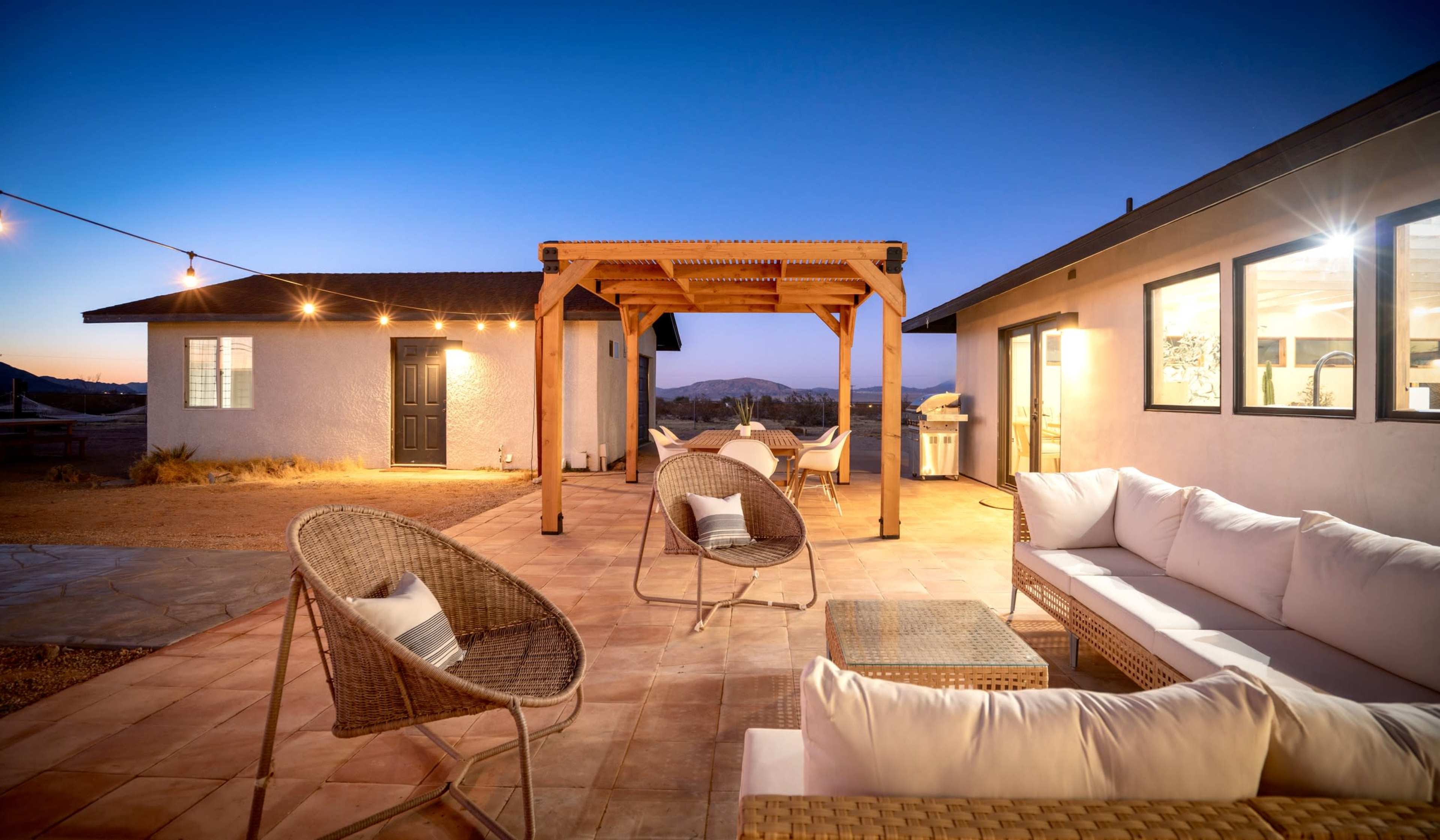 A modern patio area features seating and a pergola, illuminated by string lights at dusk.
