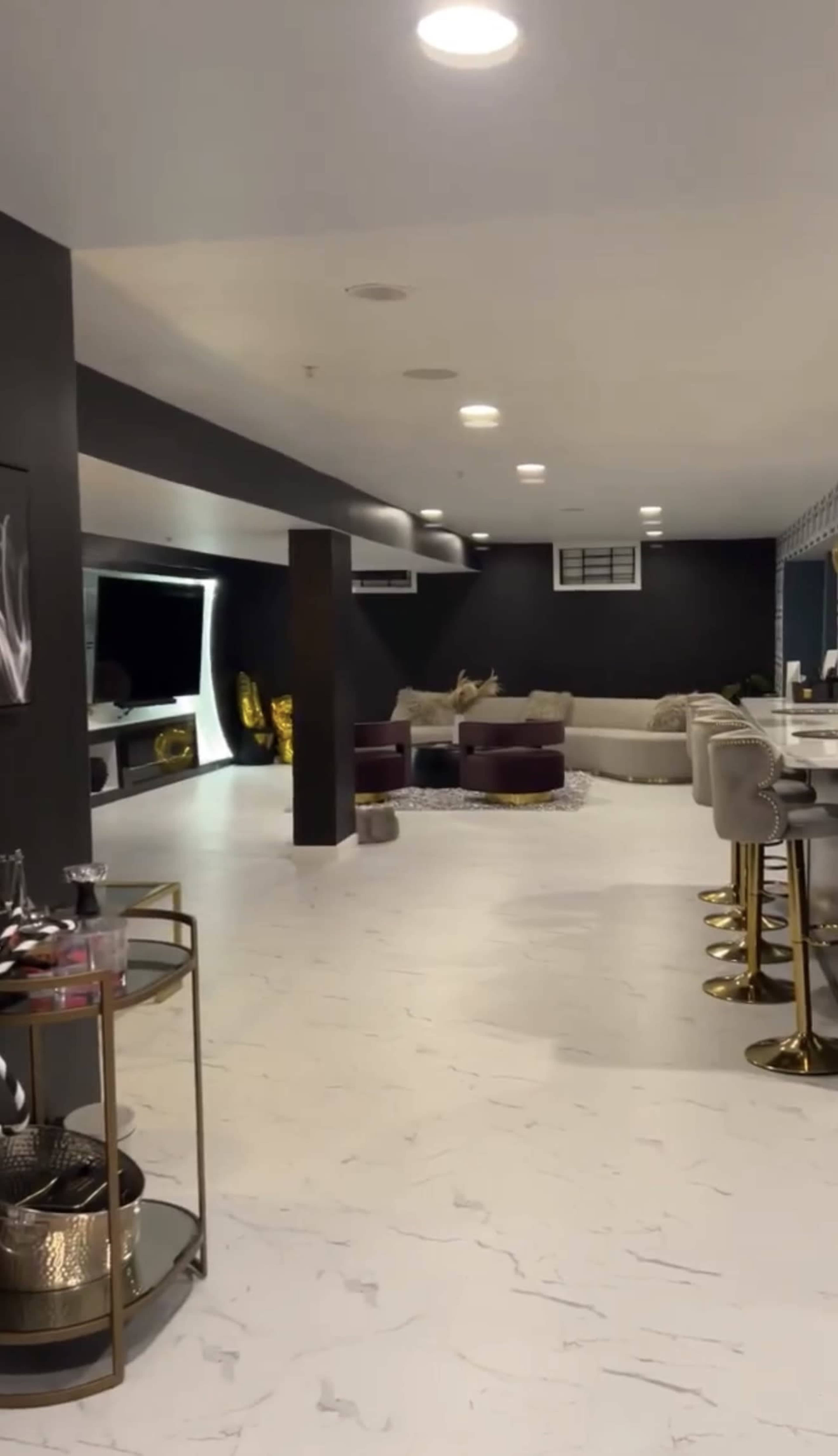 The image shows a modern, spacious living area featuring a light-colored floor, a U-shaped sectional sofa, bar stools, and large windows with dark walls.