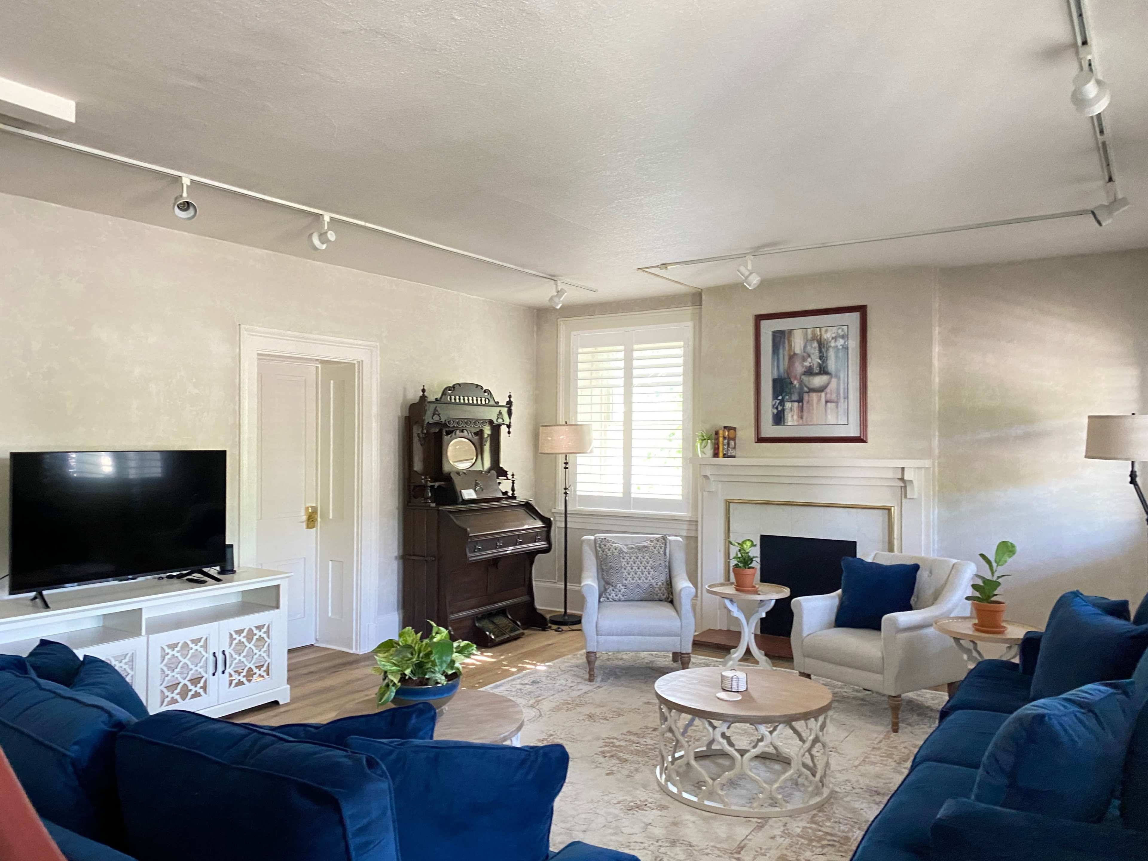 A living room features blue sofas, a television, an antique piano, and a circular coffee table, arranged around a fireplace.