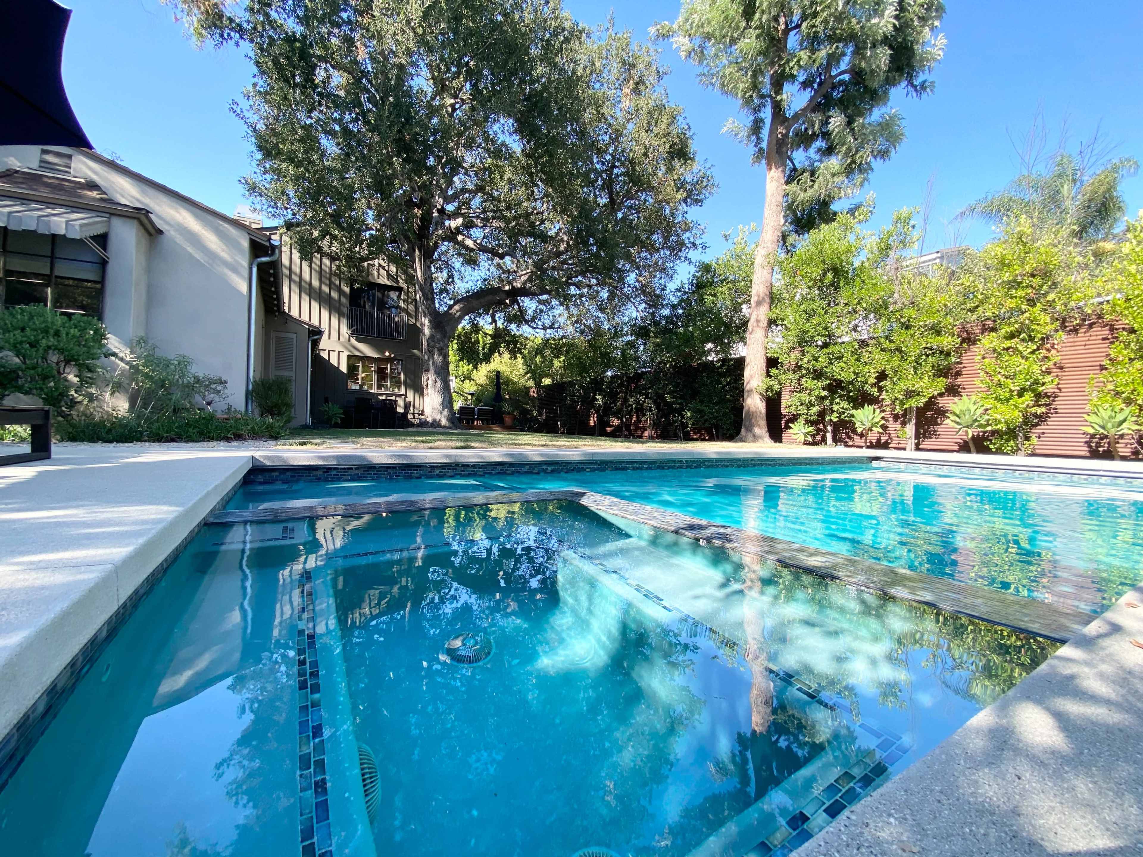 The image shows a clear swimming pool surrounded by greenery and tall trees, with a house visible in the background.