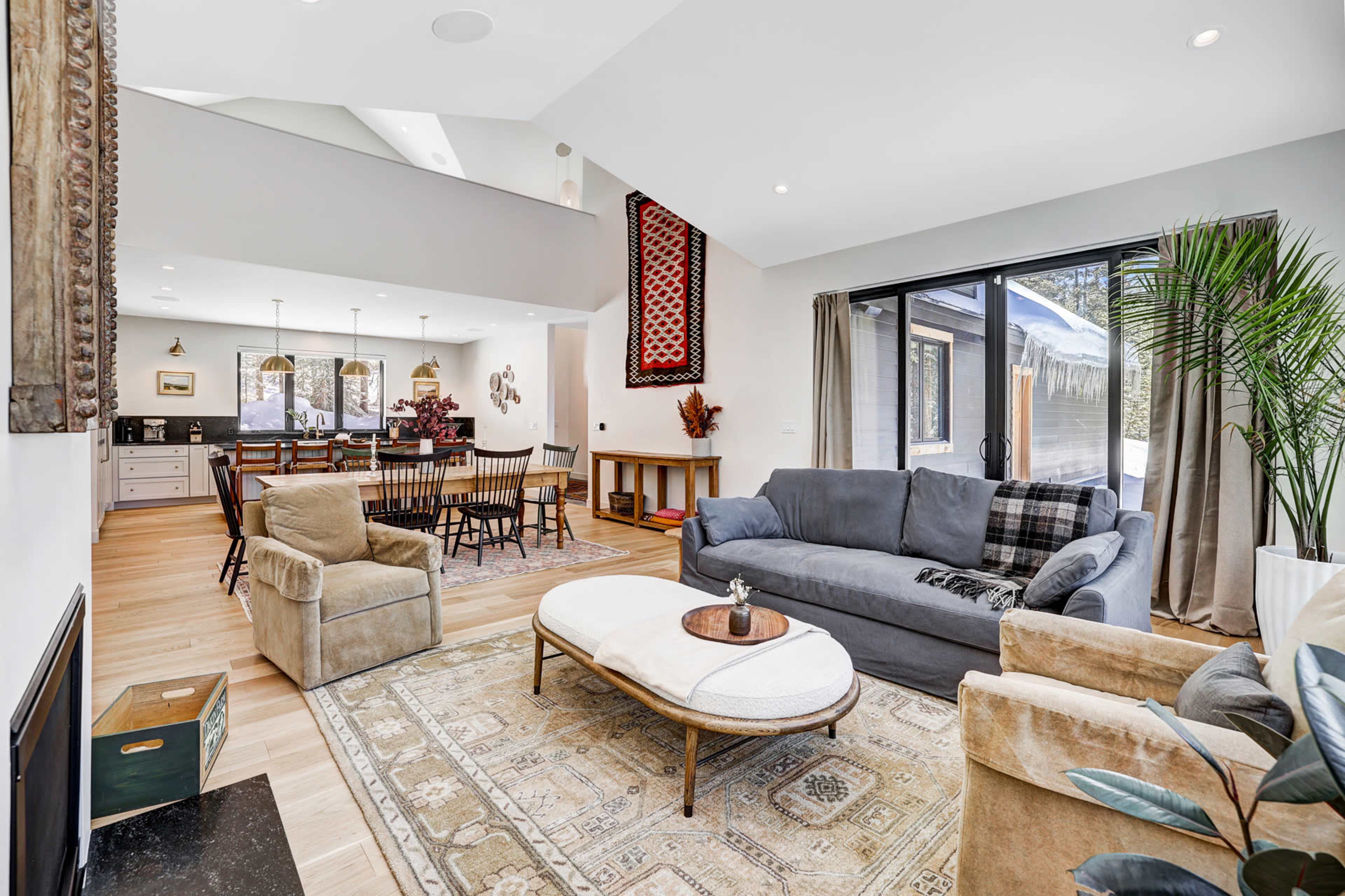 A modern living room with a gray sofa, a round coffee table, and large windows providing natural light, open to a dining area in the background.