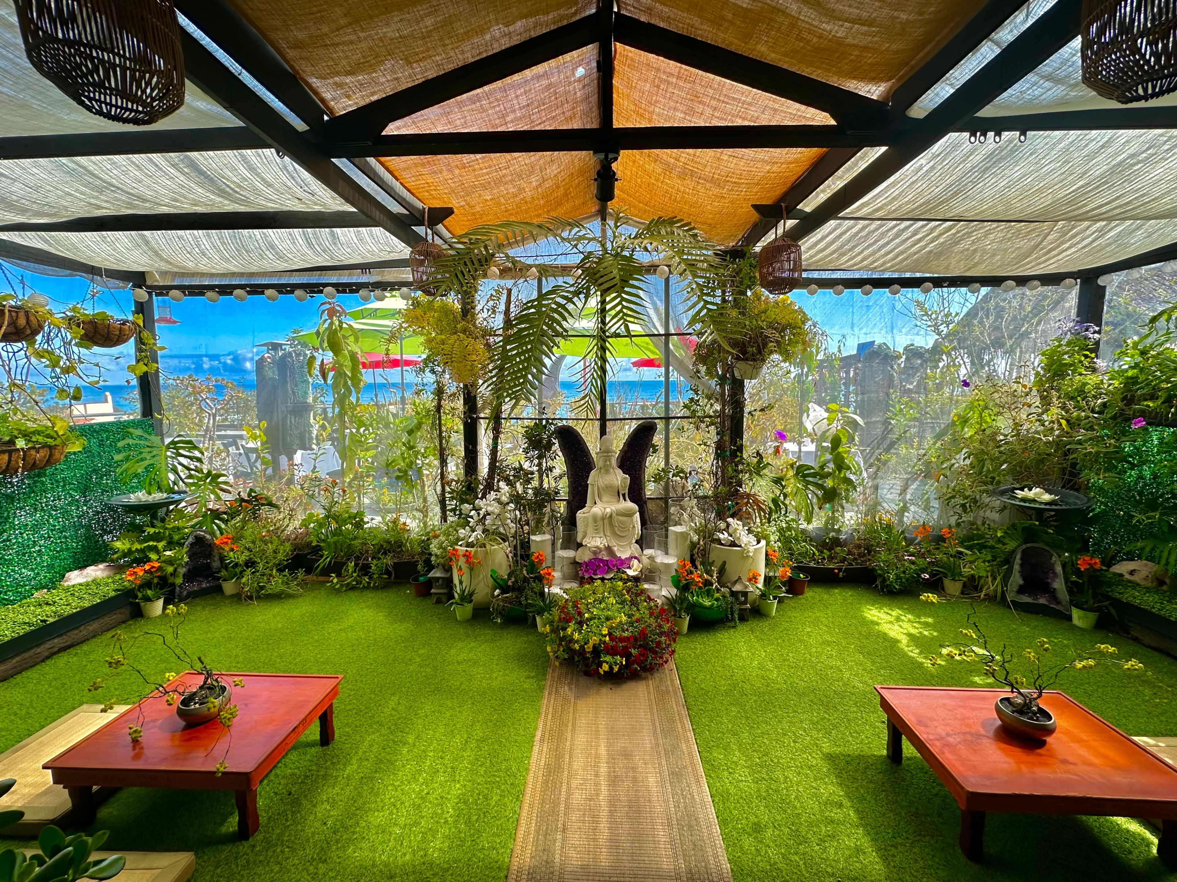 The scene depicts a serene indoor garden with green artificial turf, two wooden tables, a central Buddha statue surrounded by flowers, and abundant greenery under a canopy.