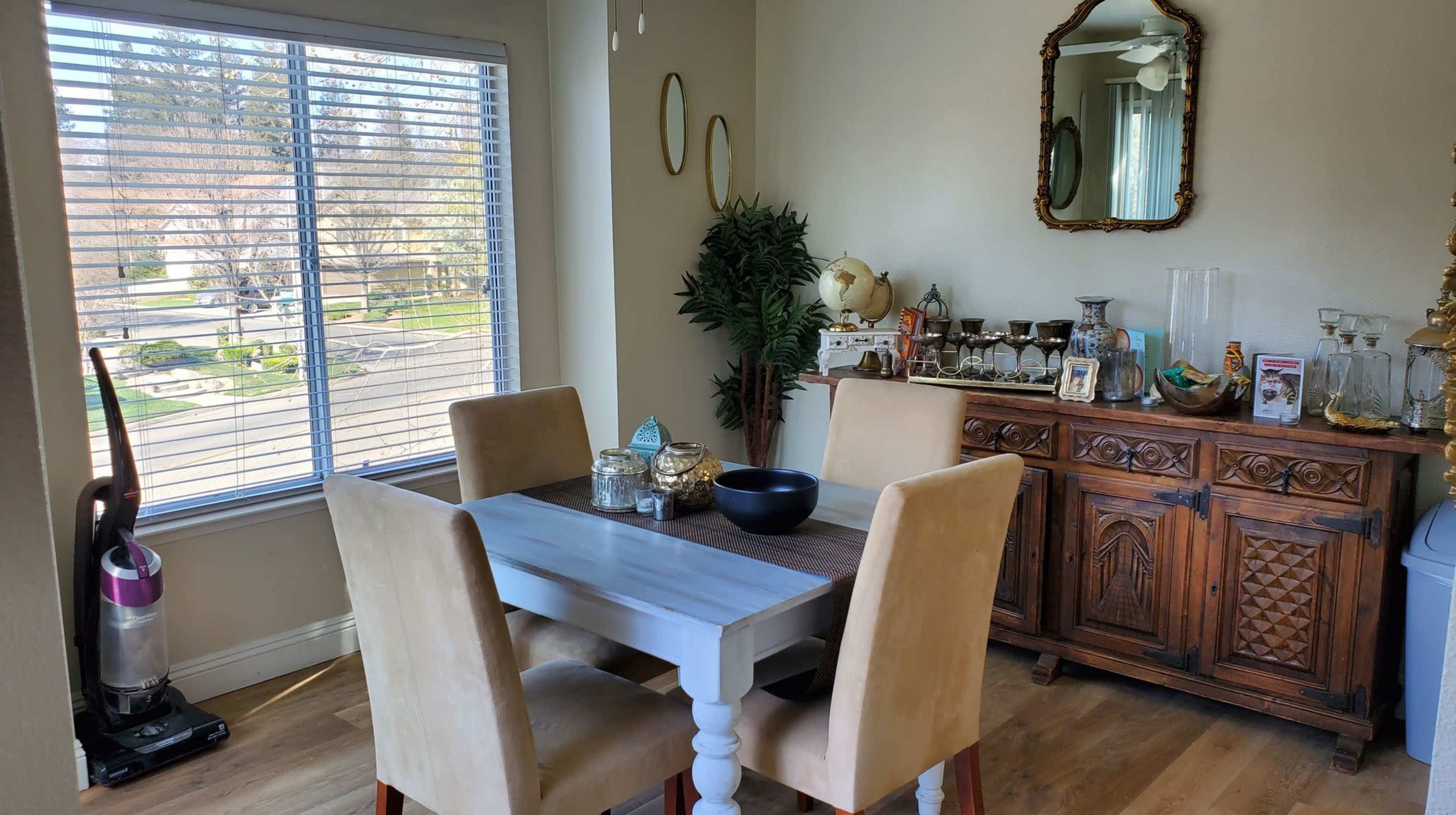 A dining area features a wooden table with four chairs, a sideboard with decorative items, and a window overlooking a street with trees.