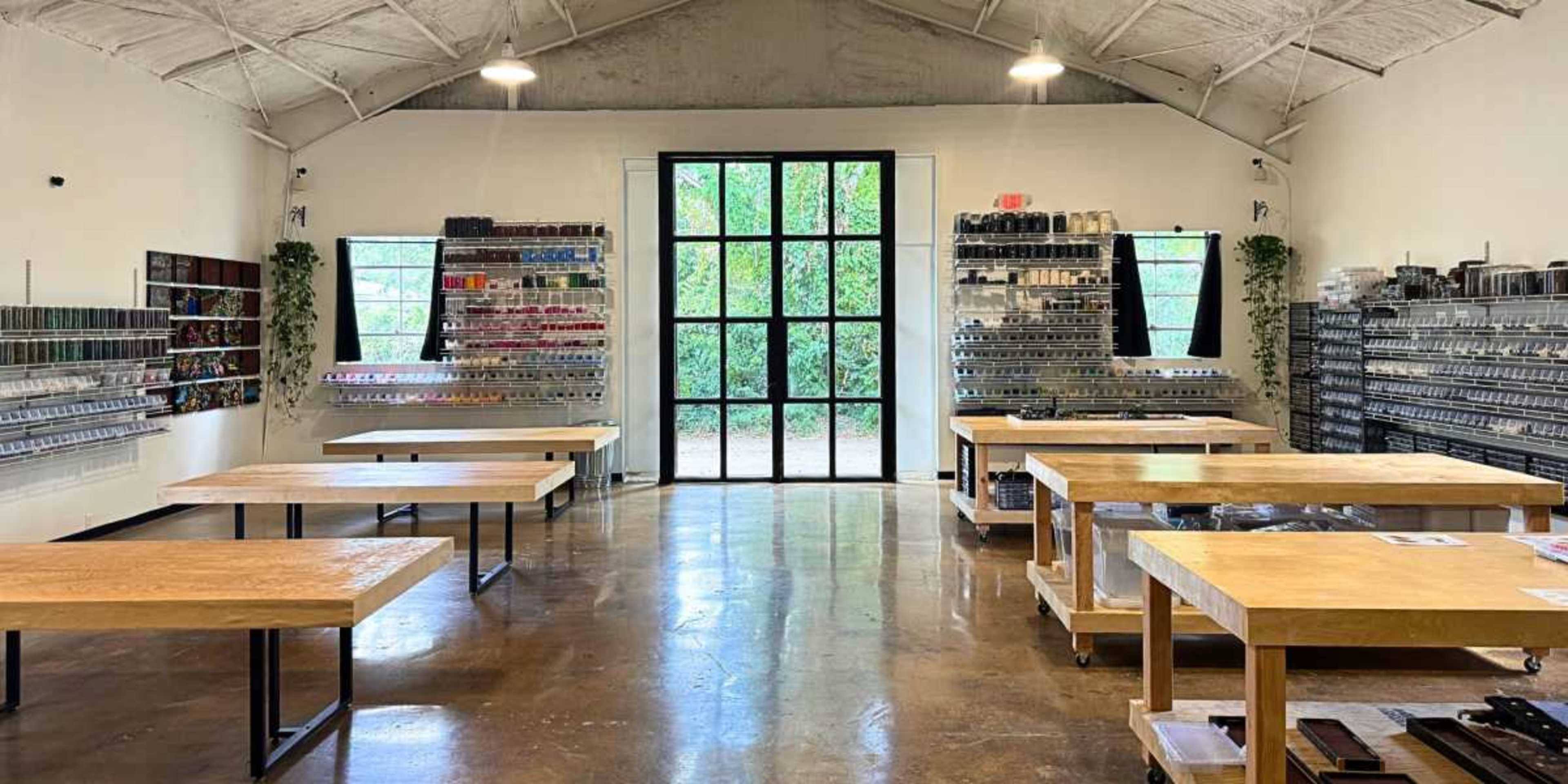The image shows a spacious, well-lit room with wooden tables and wall-mounted shelves filled with various craft supplies.