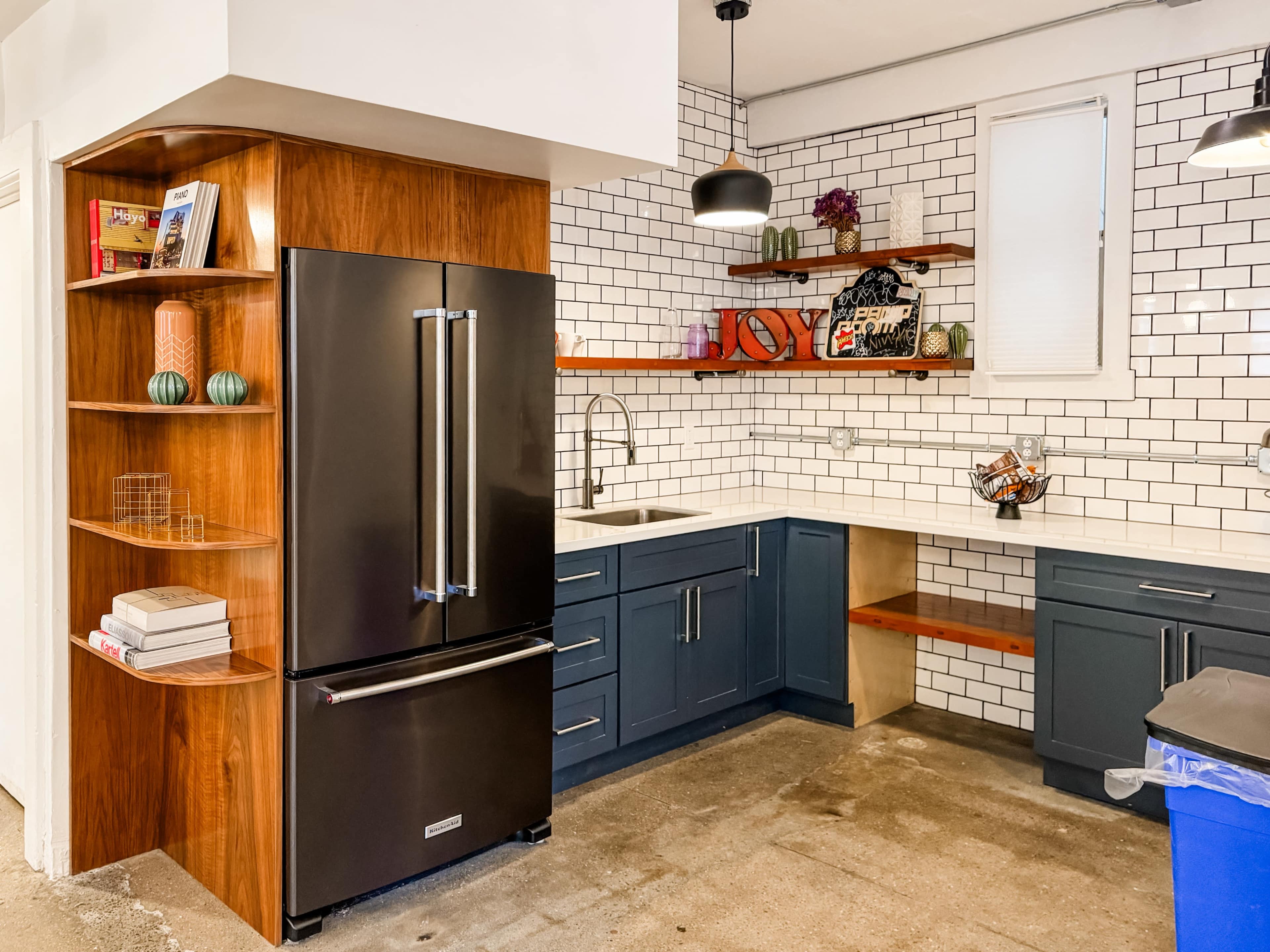 A modern kitchen featuring a black refrigerator, wooden shelving, gray cabinets, a white countertop, and subway tile backsplash.