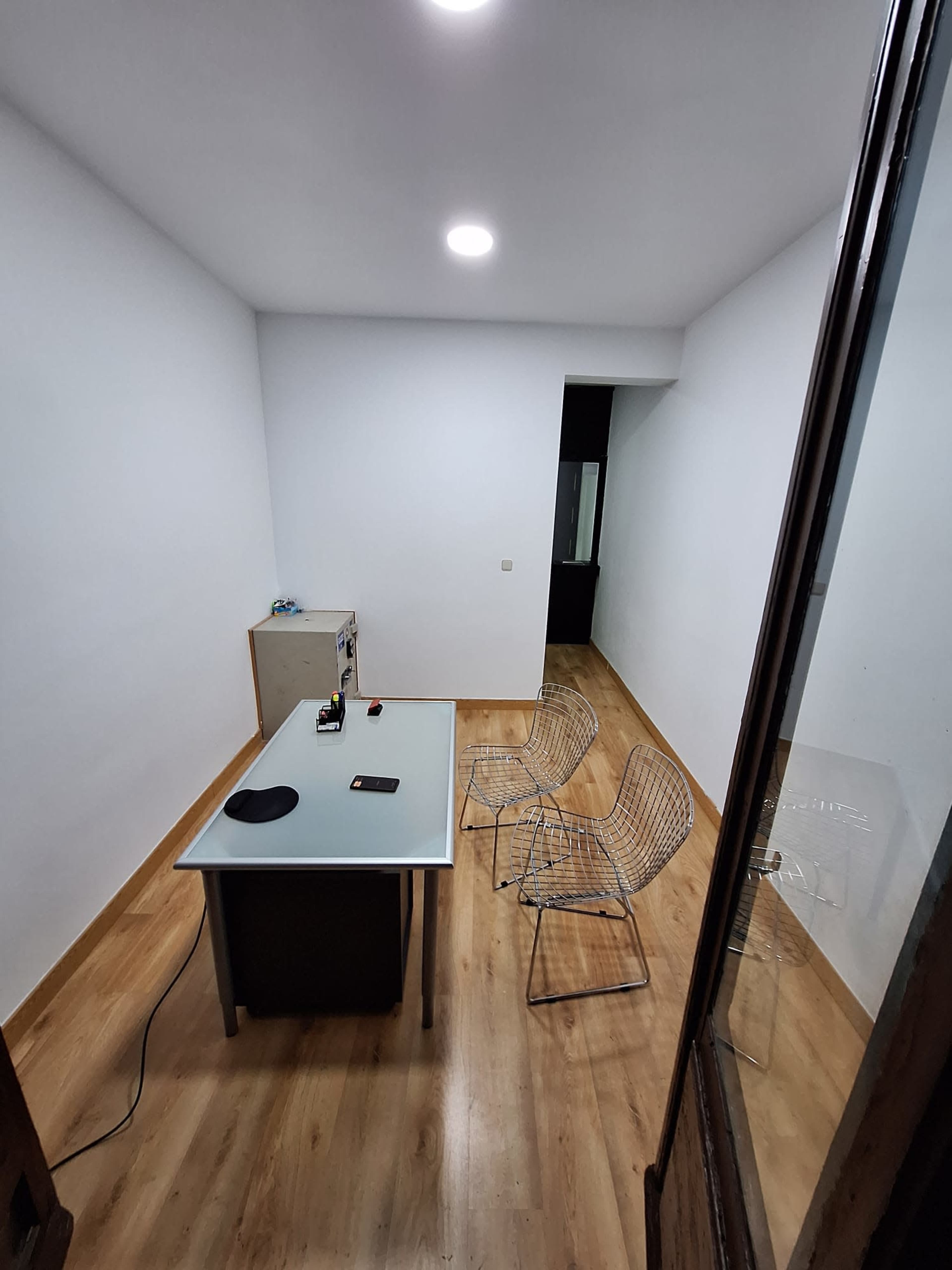 The image shows a minimalist office space with a table and two wire chairs on a wooden floor.