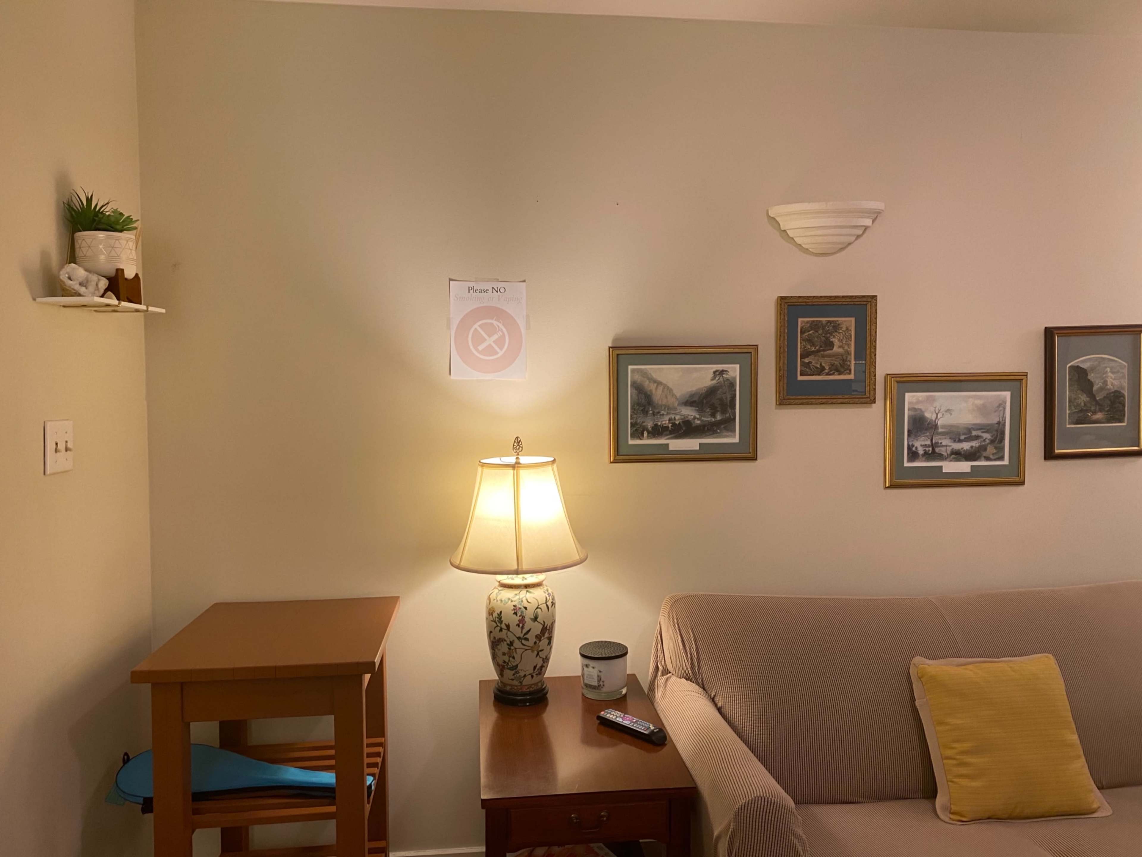 A lamp sits on a table beside a beige couch, with framed pictures hanging on the wall and a "Please No" sign displayed above.