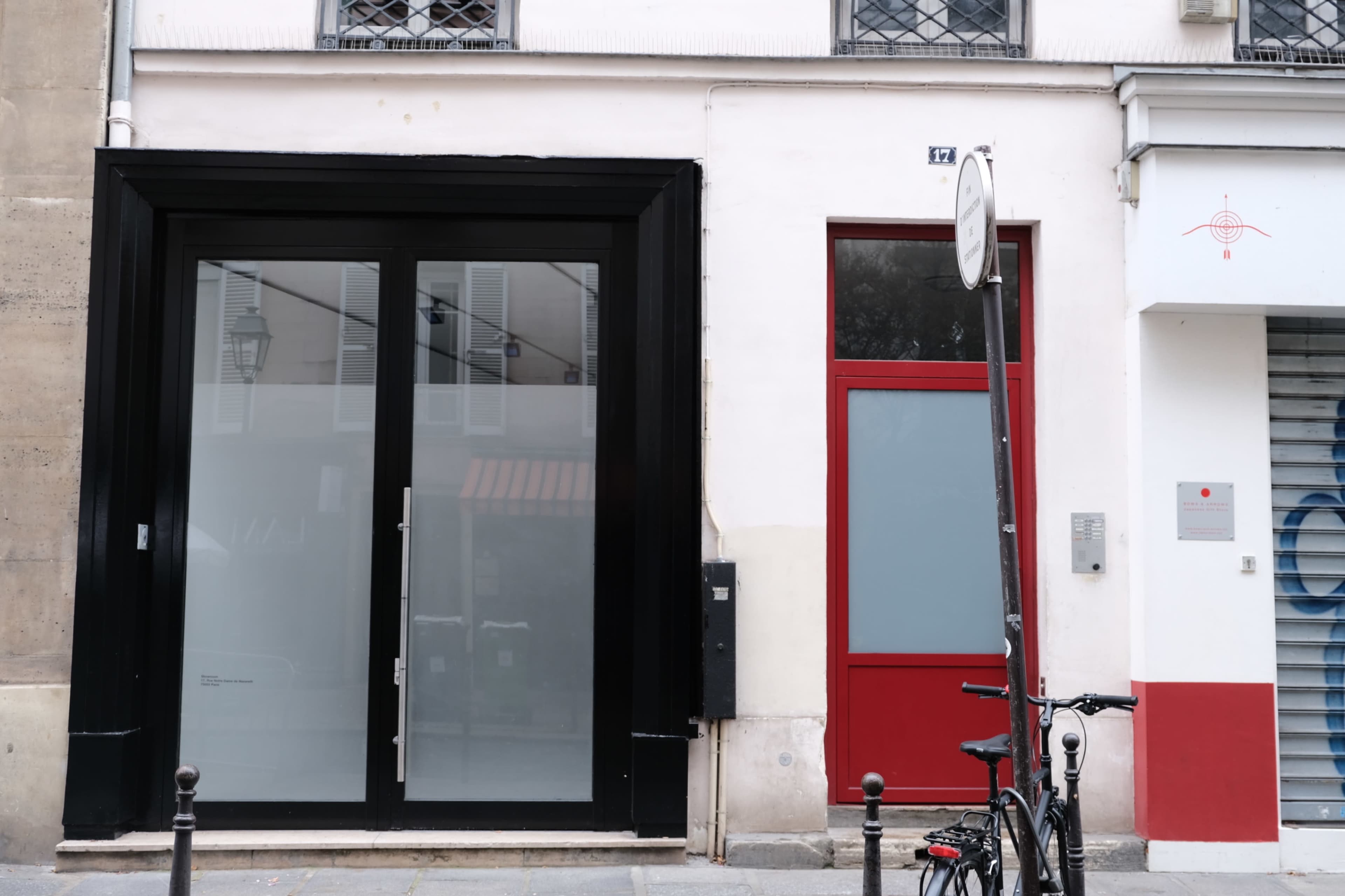 The image shows two adjacent storefronts, one with a large black glass facade and the other with a red door, alongside a parked bicycle and a street sign.