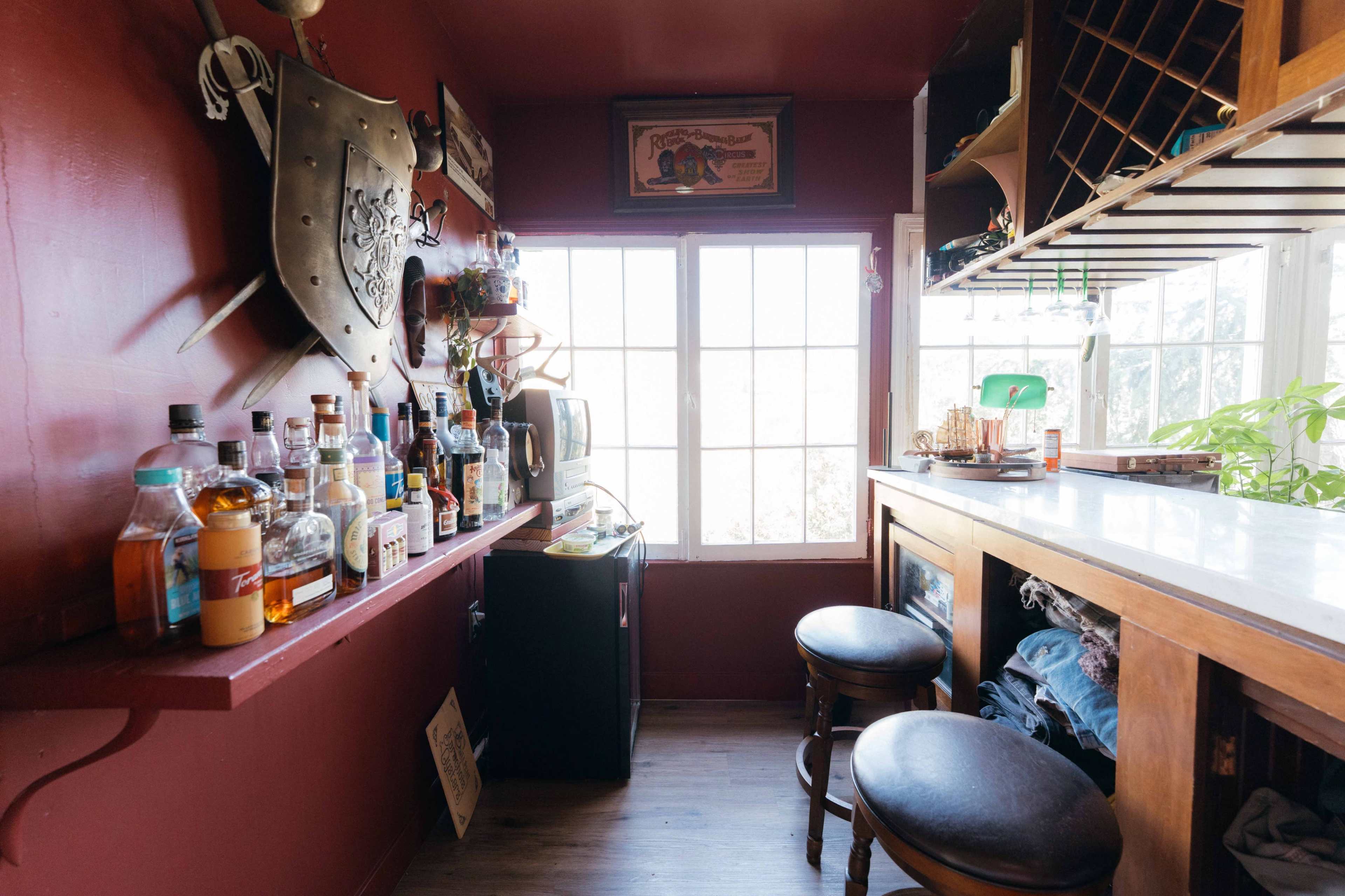 The image shows a small, dimly lit bar area with a wooden counter, a collection of bottles on a shelf, and a window allowing natural light to enter.