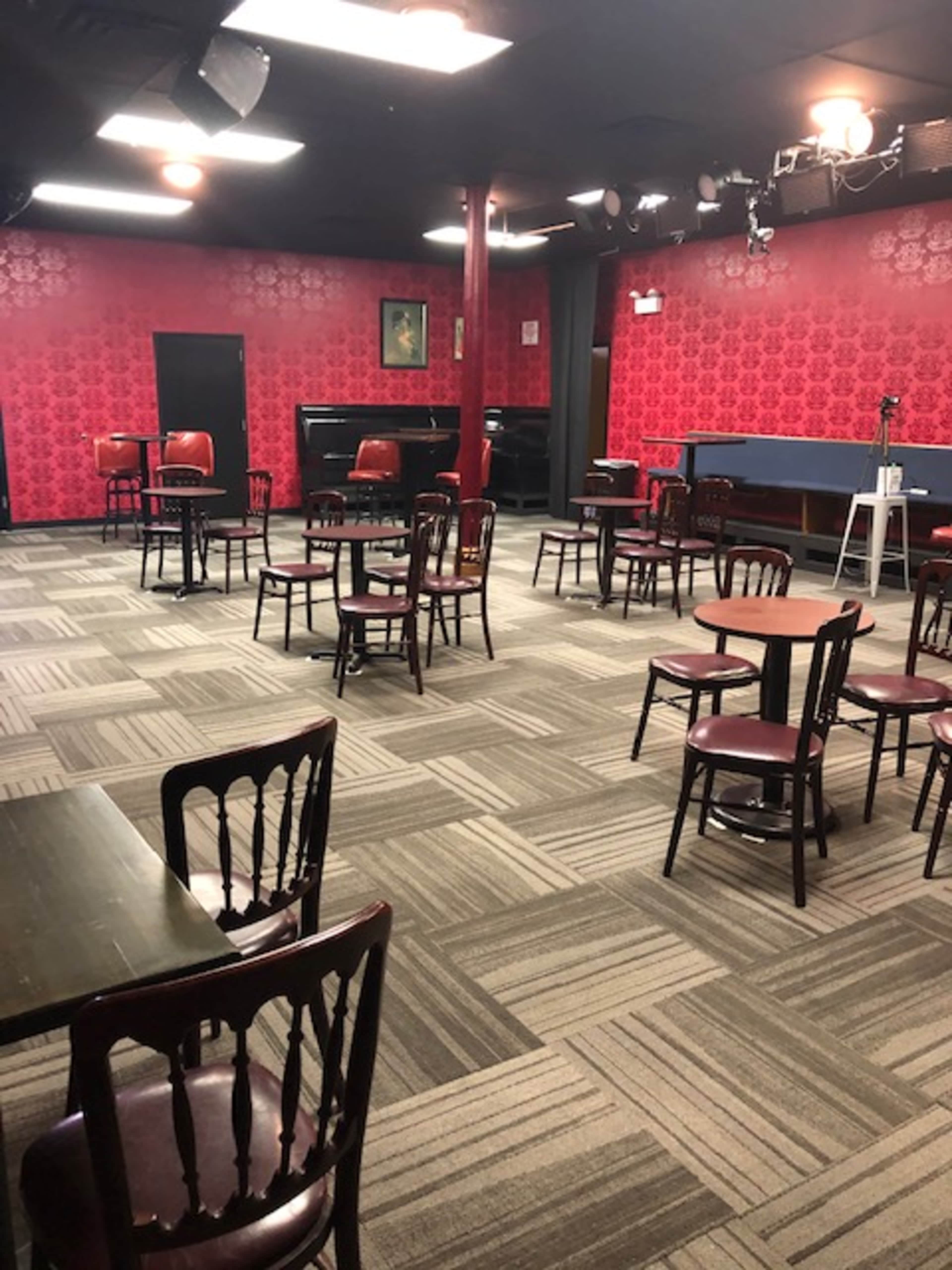 An empty performance venue with red patterned walls, several chairs scattered around tables, and a small stage area at the front.