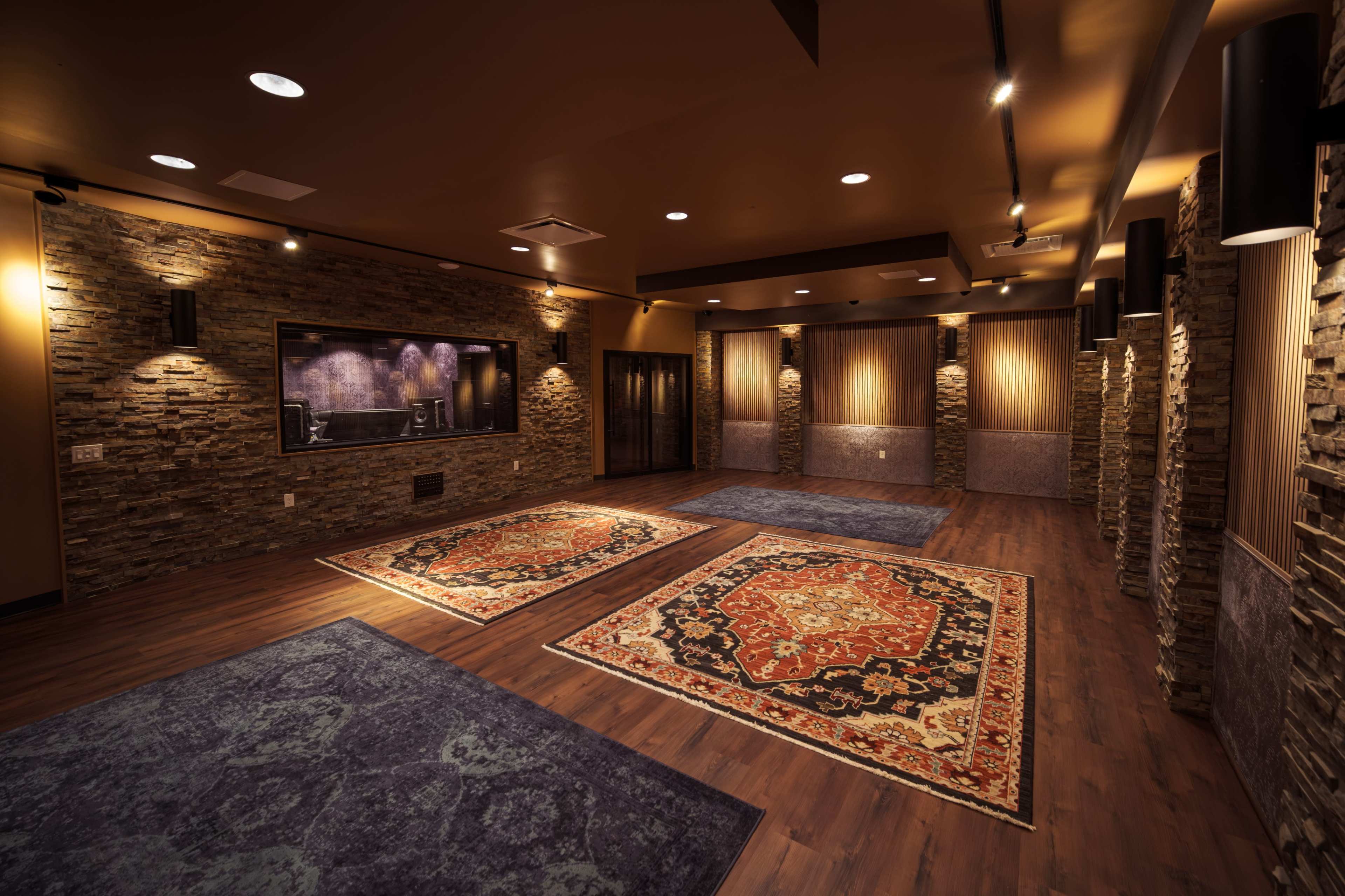 The image shows a spacious, well-lit interior with stone walls and wooden flooring, featuring large area rugs and a sound stage area.