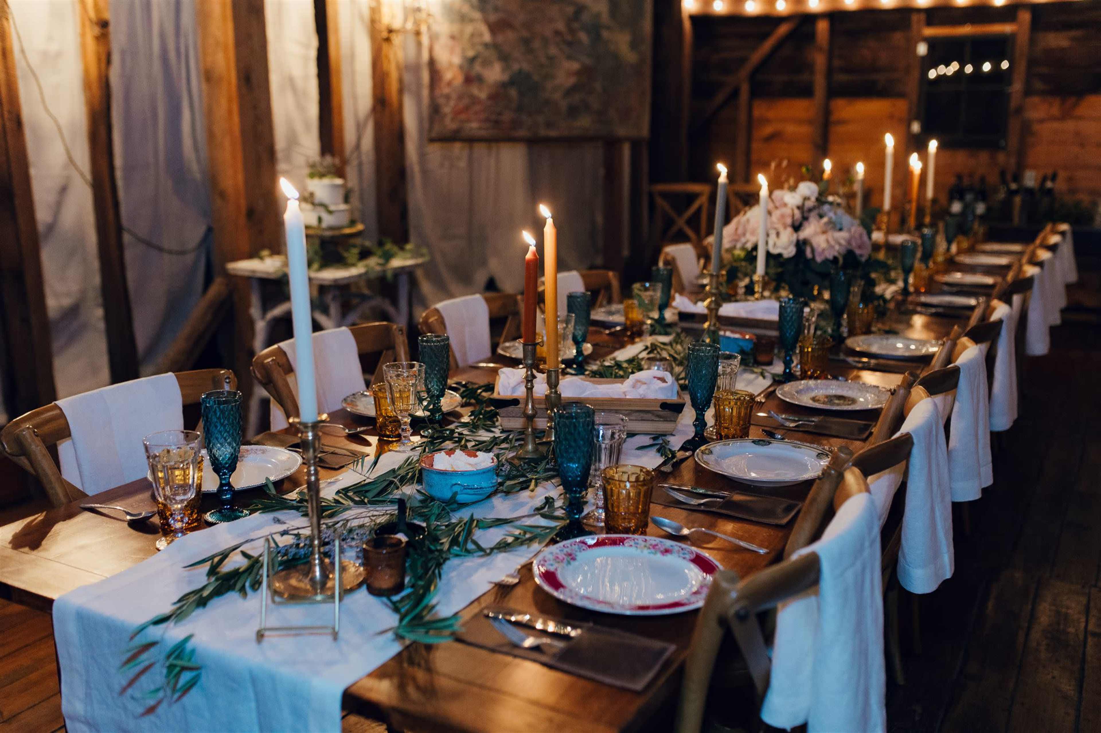 A long dining table is elegantly set with candles, glassware, and floral arrangements in a rustic indoor space.