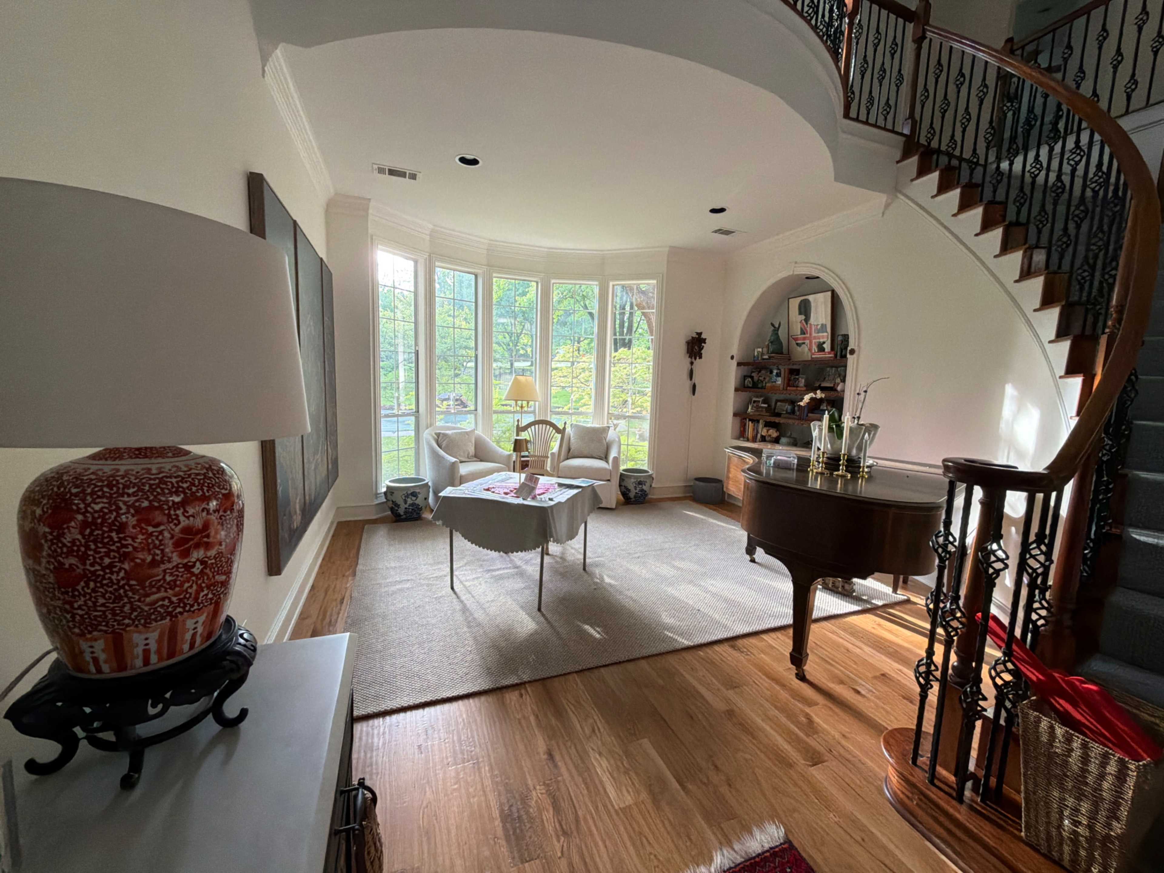 The image shows a well-lit entryway with a curved staircase and a seating area featuring two chairs and a small table beside a large window.
