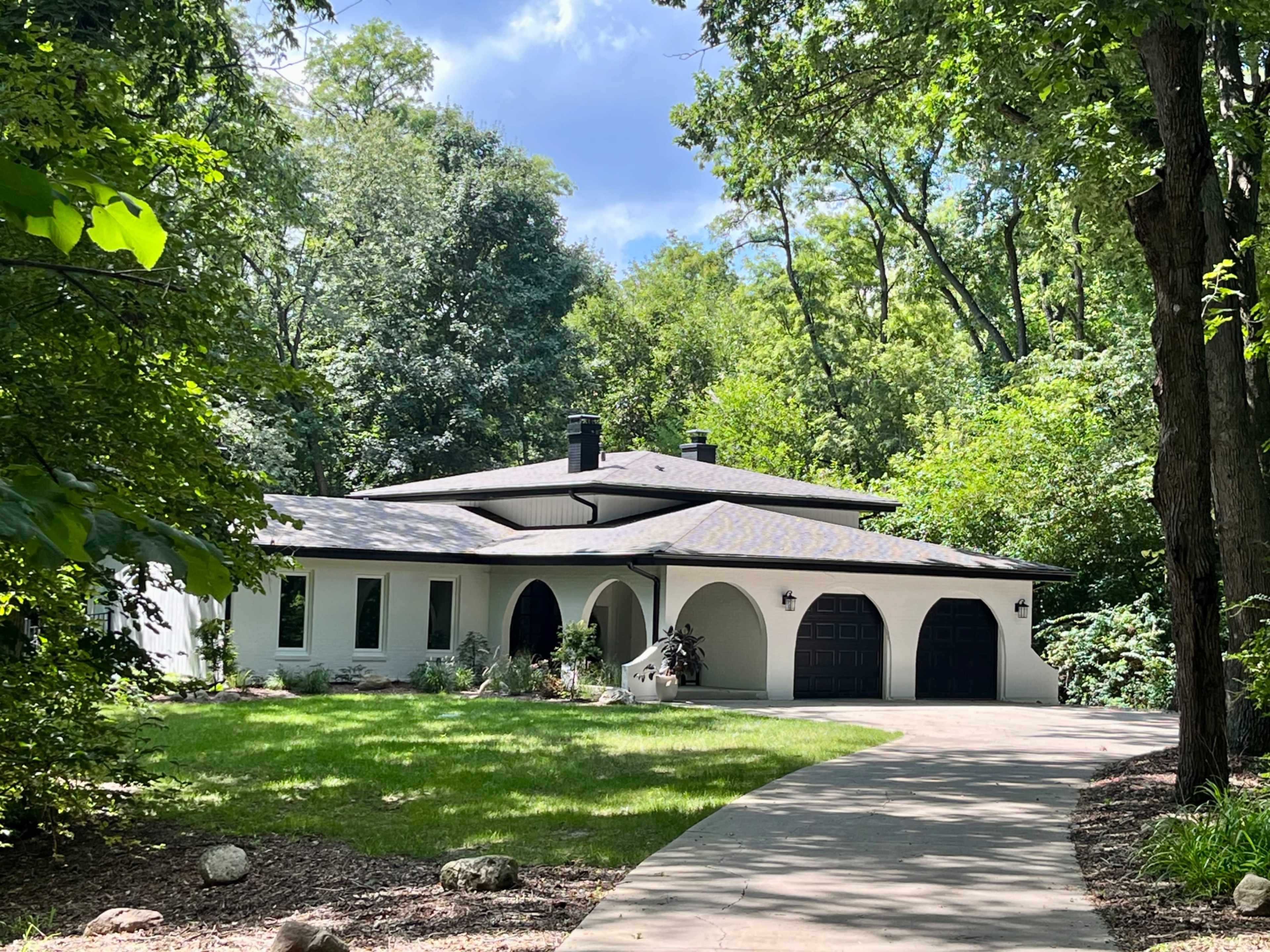 A modern, single-story house with a curved roof and large windows is set amidst lush greenery, with a winding driveway leading to a double garage.