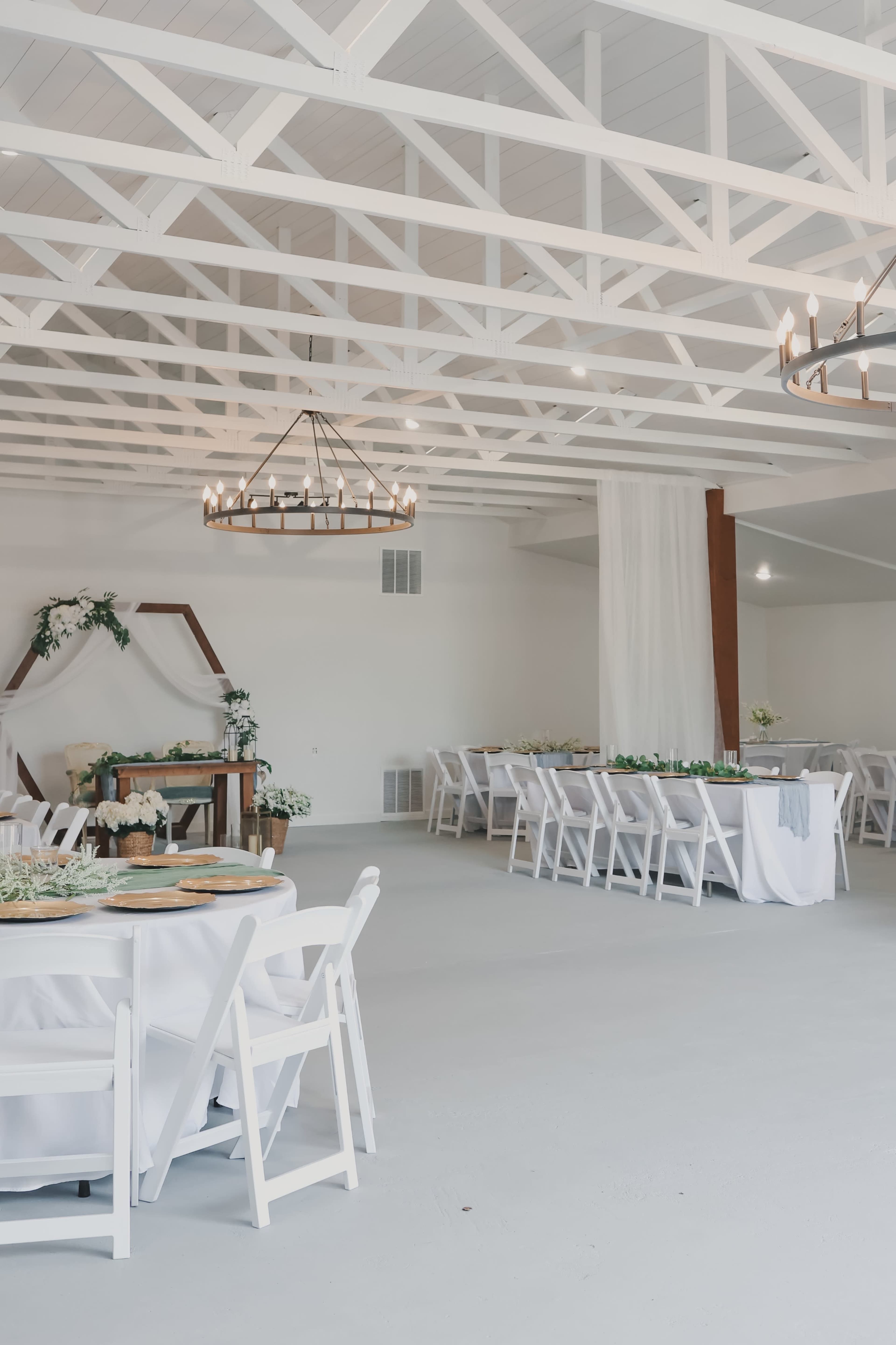 The image shows a spacious indoor venue set up for an event, featuring white tables and chairs, a wooden centerpiece table, and decorative greenery against white walls and beams.