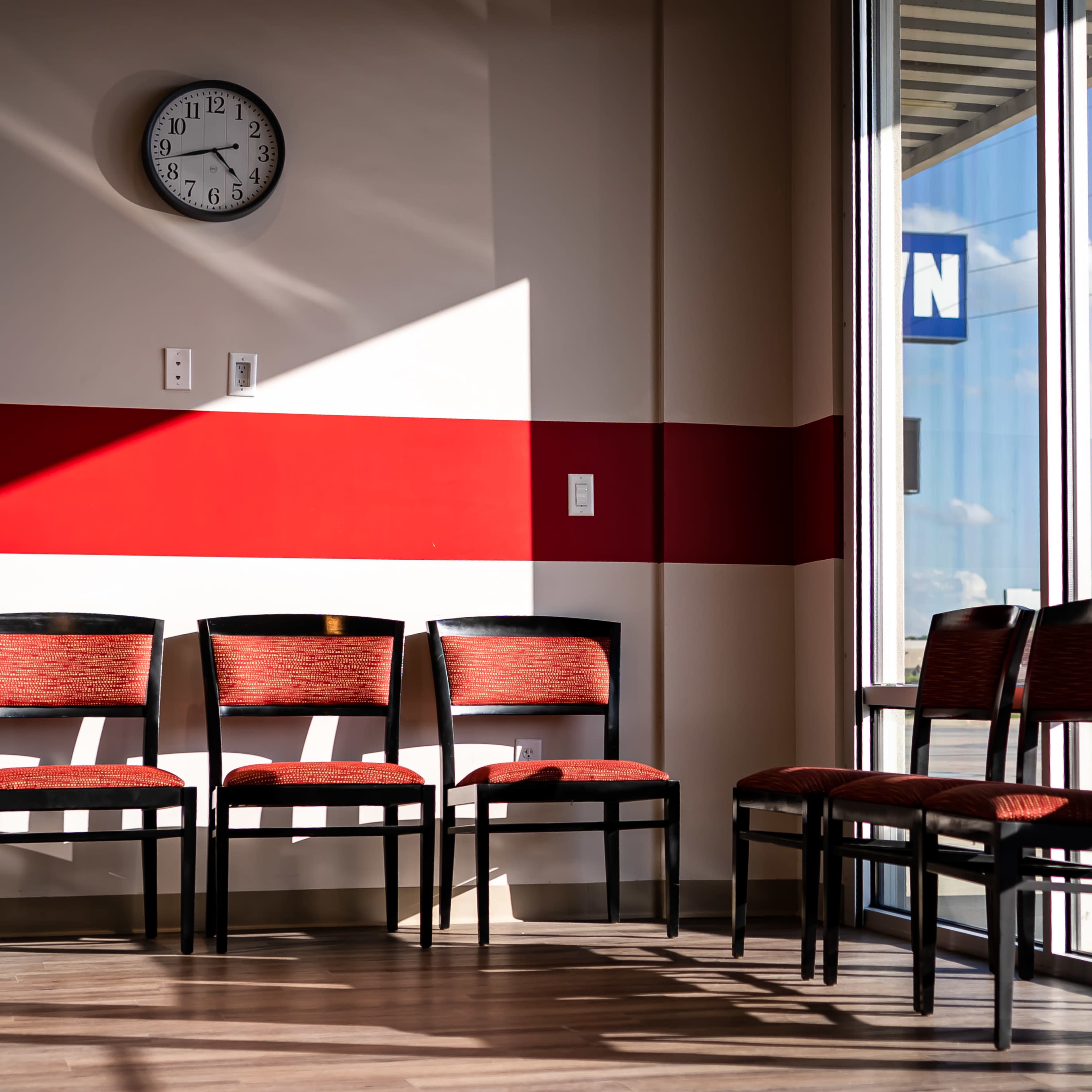 A row of six black chairs with red upholstery positioned against a wall, illuminated by sunlight streaming through a large window.