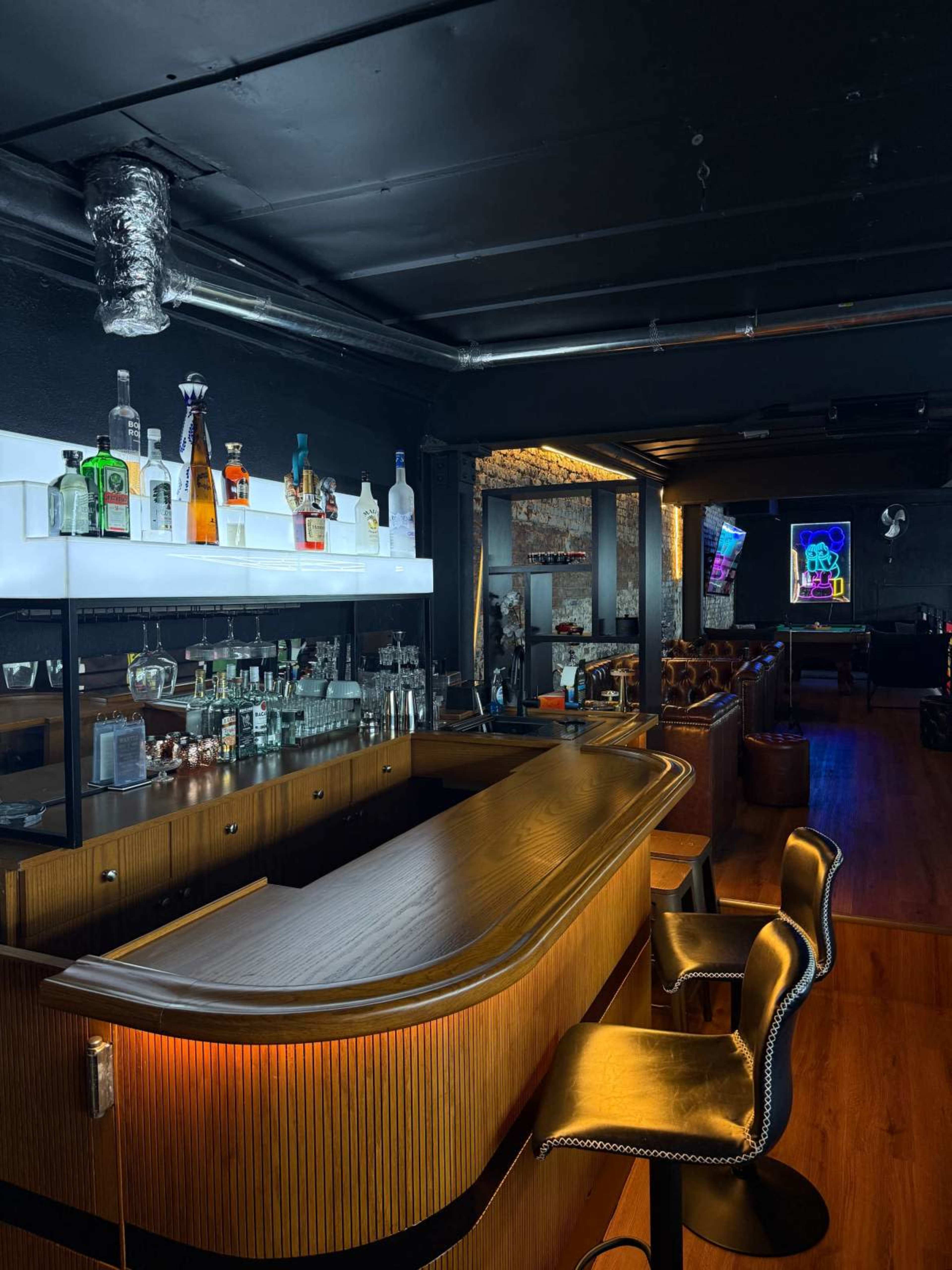 The image shows a dimly lit bar area with a polished wooden counter, shelves lined with various liquor bottles, and leather stools.