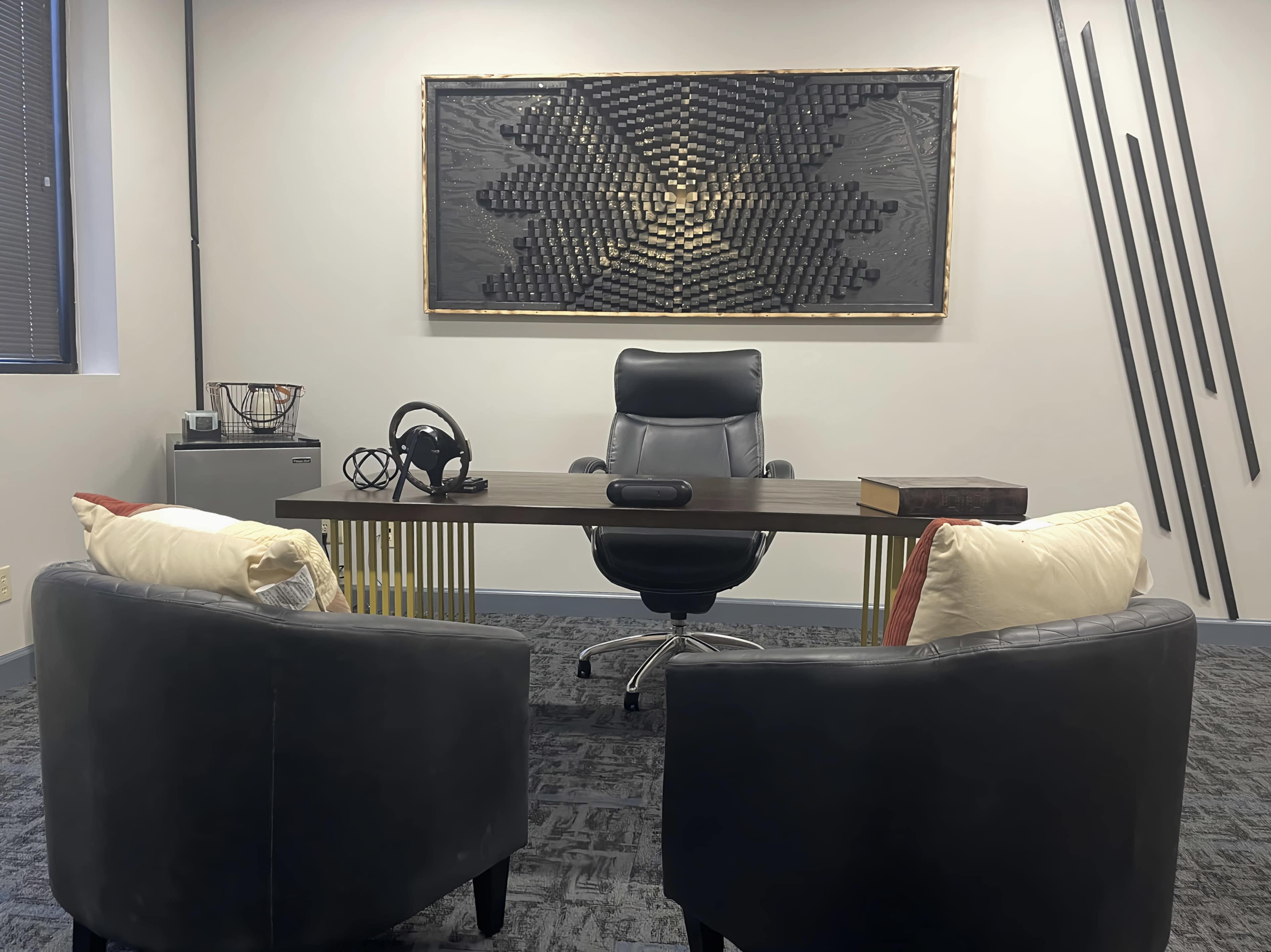 A modern office space with a desk, an ergonomic chair, and two upholstered armchairs facing each other, accompanied by a decorative wall piece made of arranged objects.