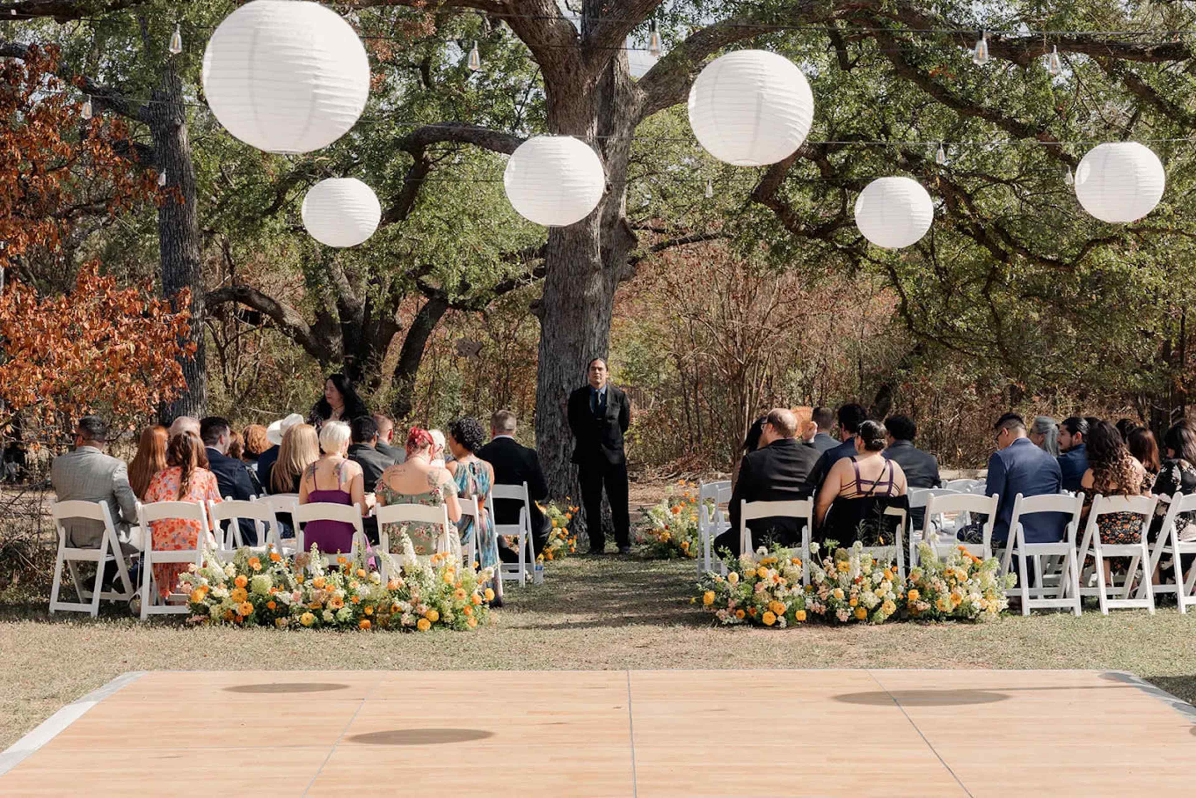 A wedding ceremony takes place outdoors with guests seated in white chairs, large lanterns hanging above, and a floral arrangement at the altar.