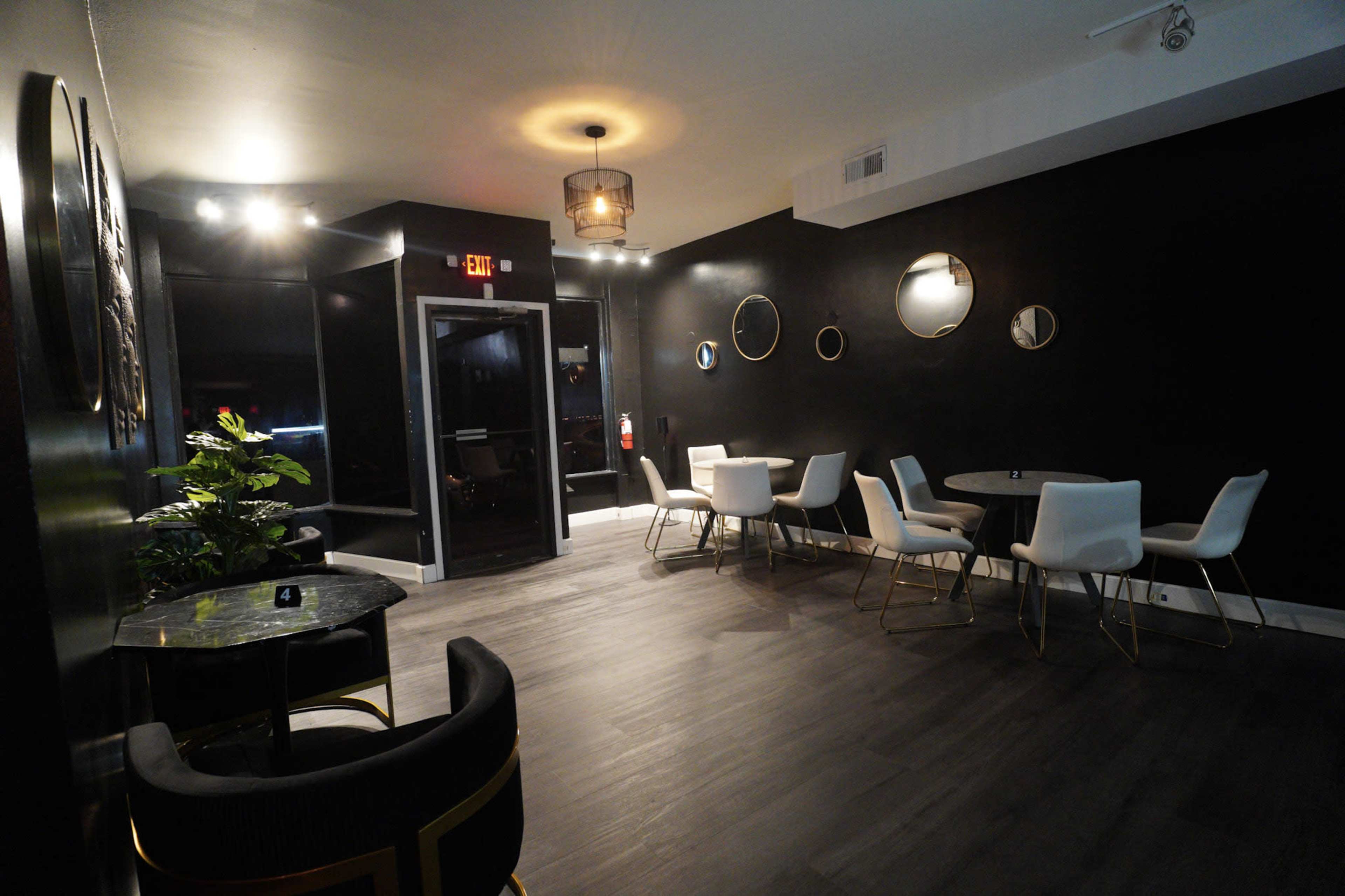 The image shows a modern, dimly lit interior of a restaurant or café featuring black walls, tables with white chairs, and decorative wall mirrors.