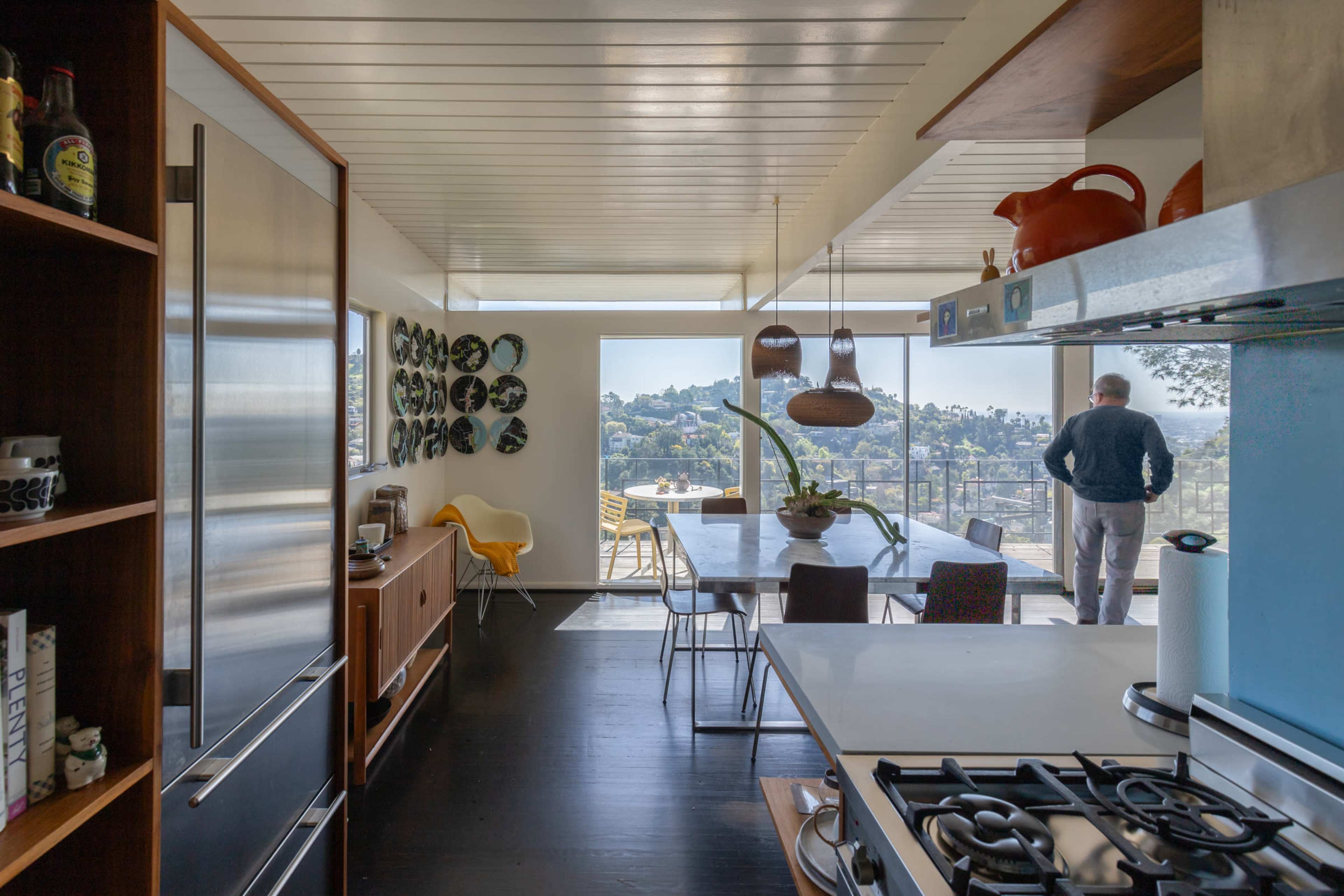 A modern kitchen opens to a dining area with a view, featuring sleek appliances, a dining table, and a man looking out through large windows.