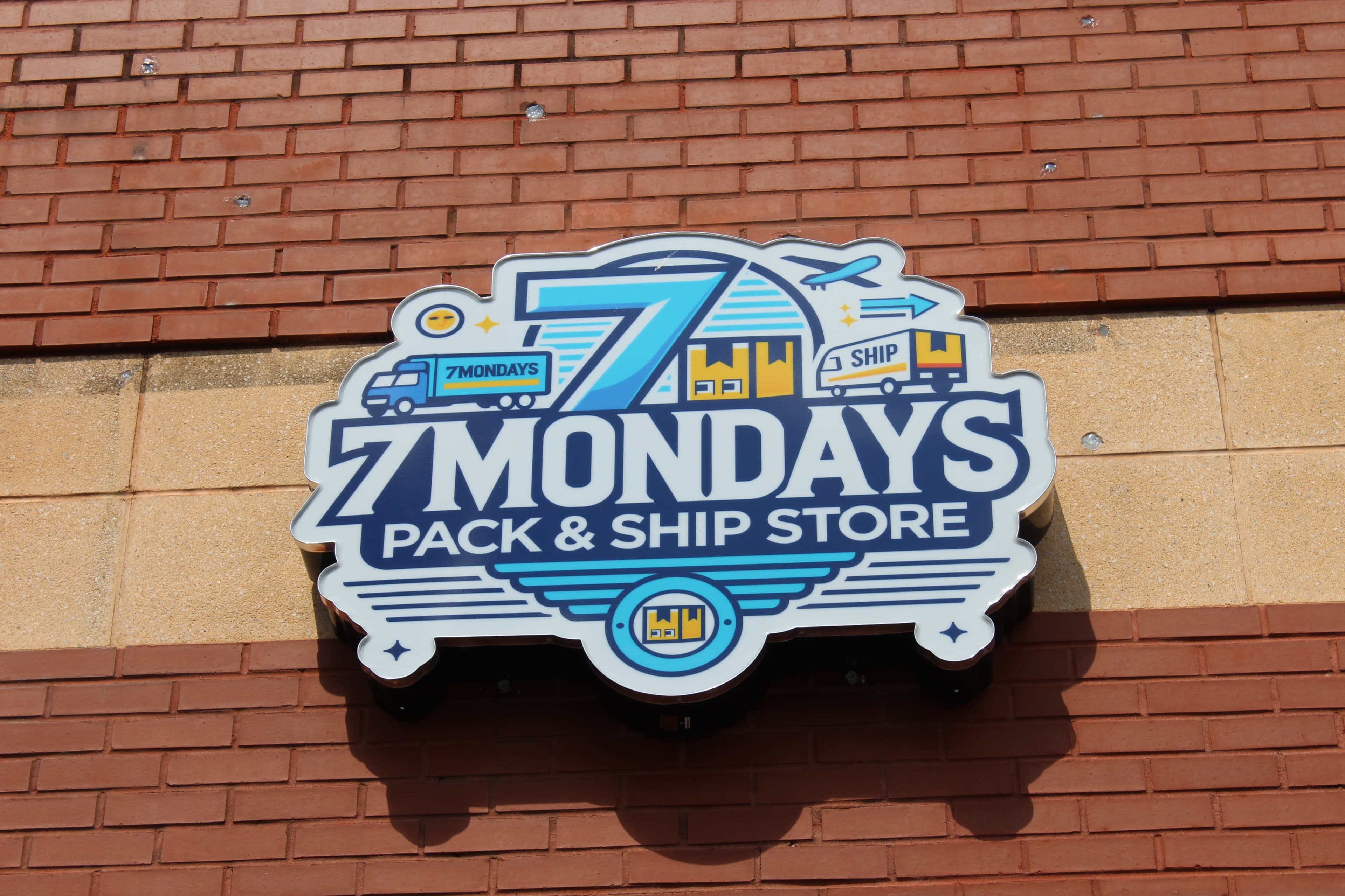 The image shows a colorful sign for "7 Mondays Pack & Ship Store," featuring imagery of transportation vehicles and a modern graphic design.