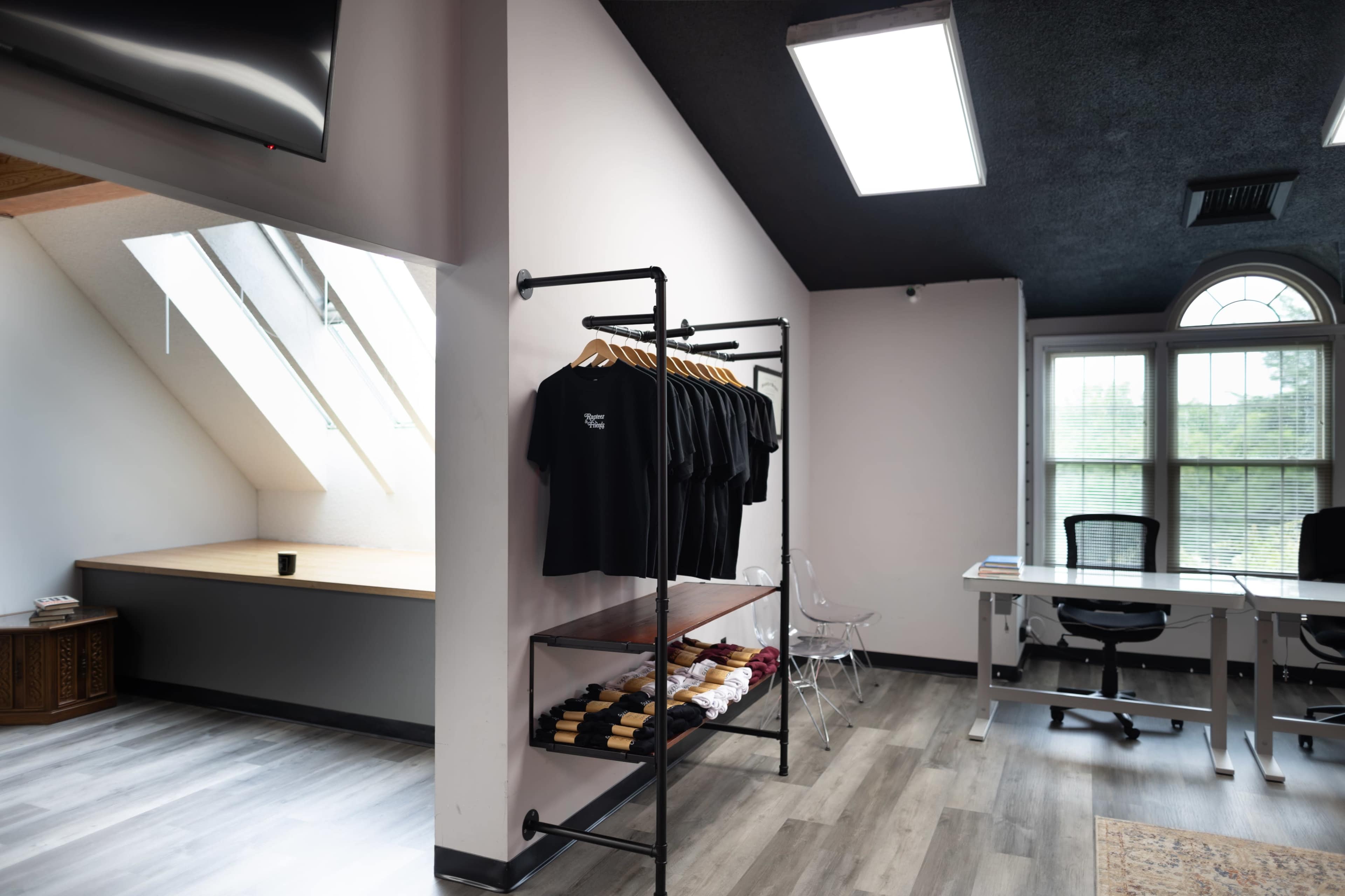 The image shows a modern interior space featuring a clothing display rack with black shirts, a wooden table, and an office desk under a sloped ceiling with skylights.