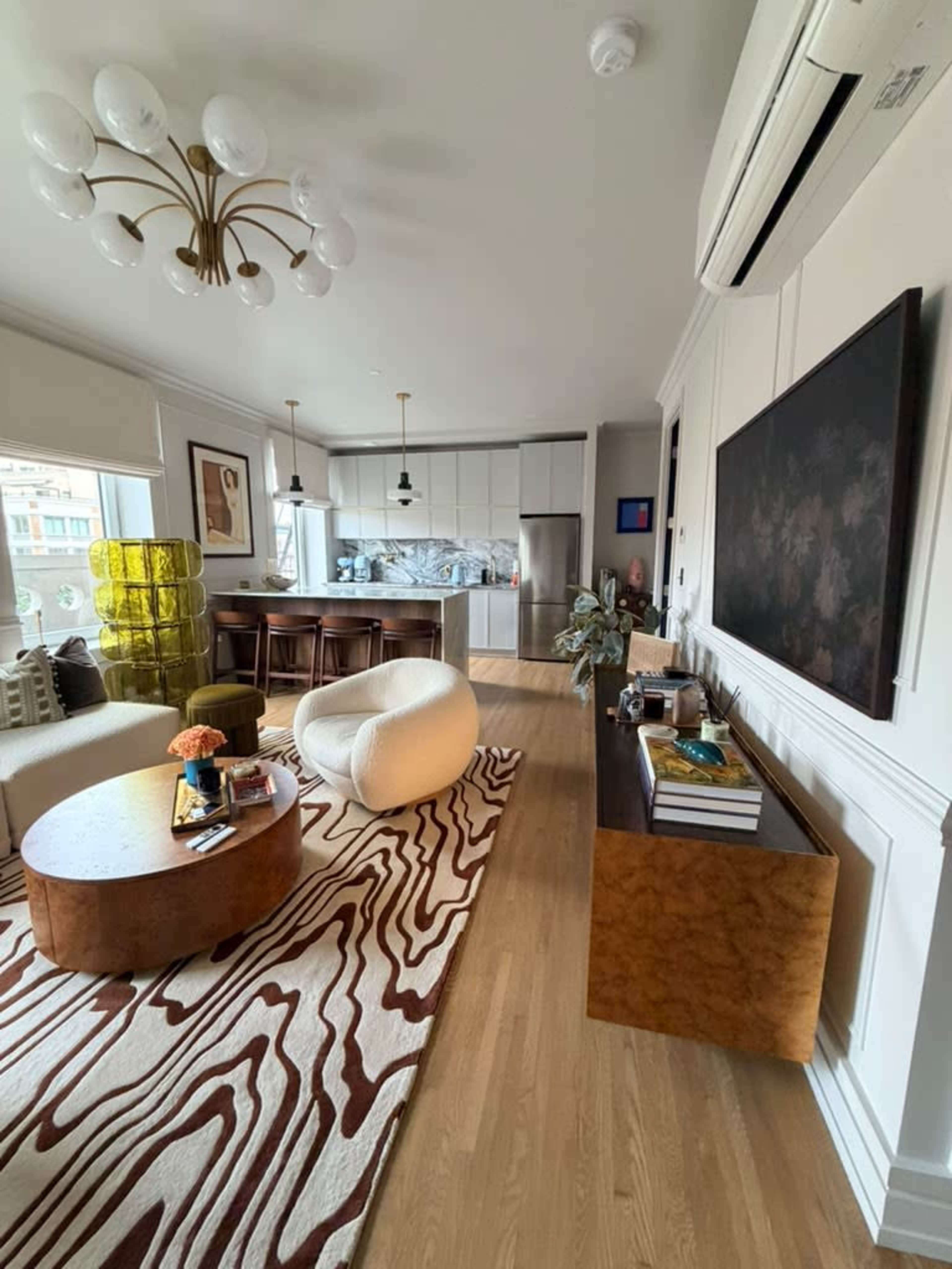 The image shows a modern living room with a circular coffee table, a patterned rug, and a kitchen area in the background.