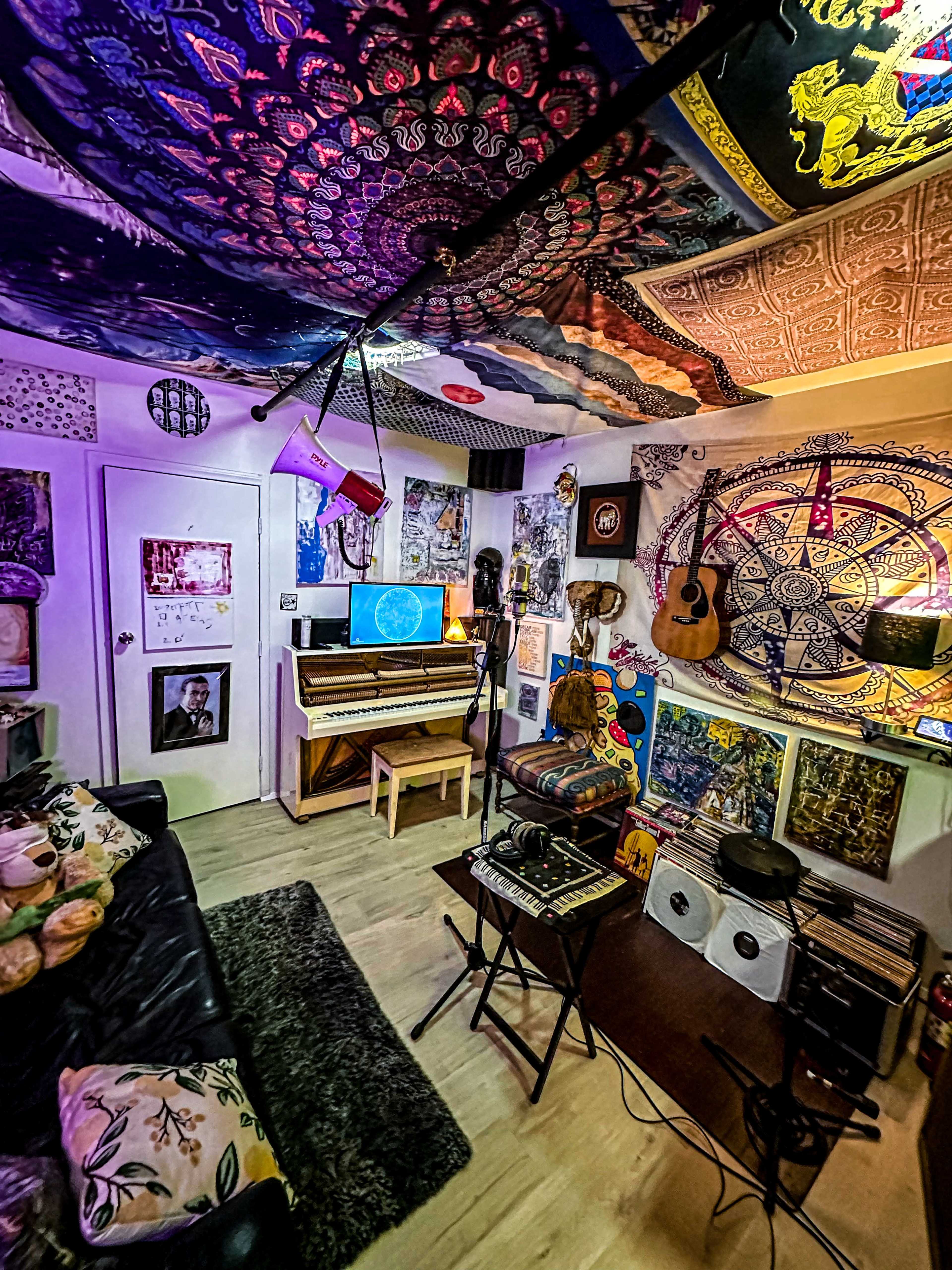 The image shows a music studio or creative space filled with musical instruments, vibrant tapestries, and various artwork on the walls.