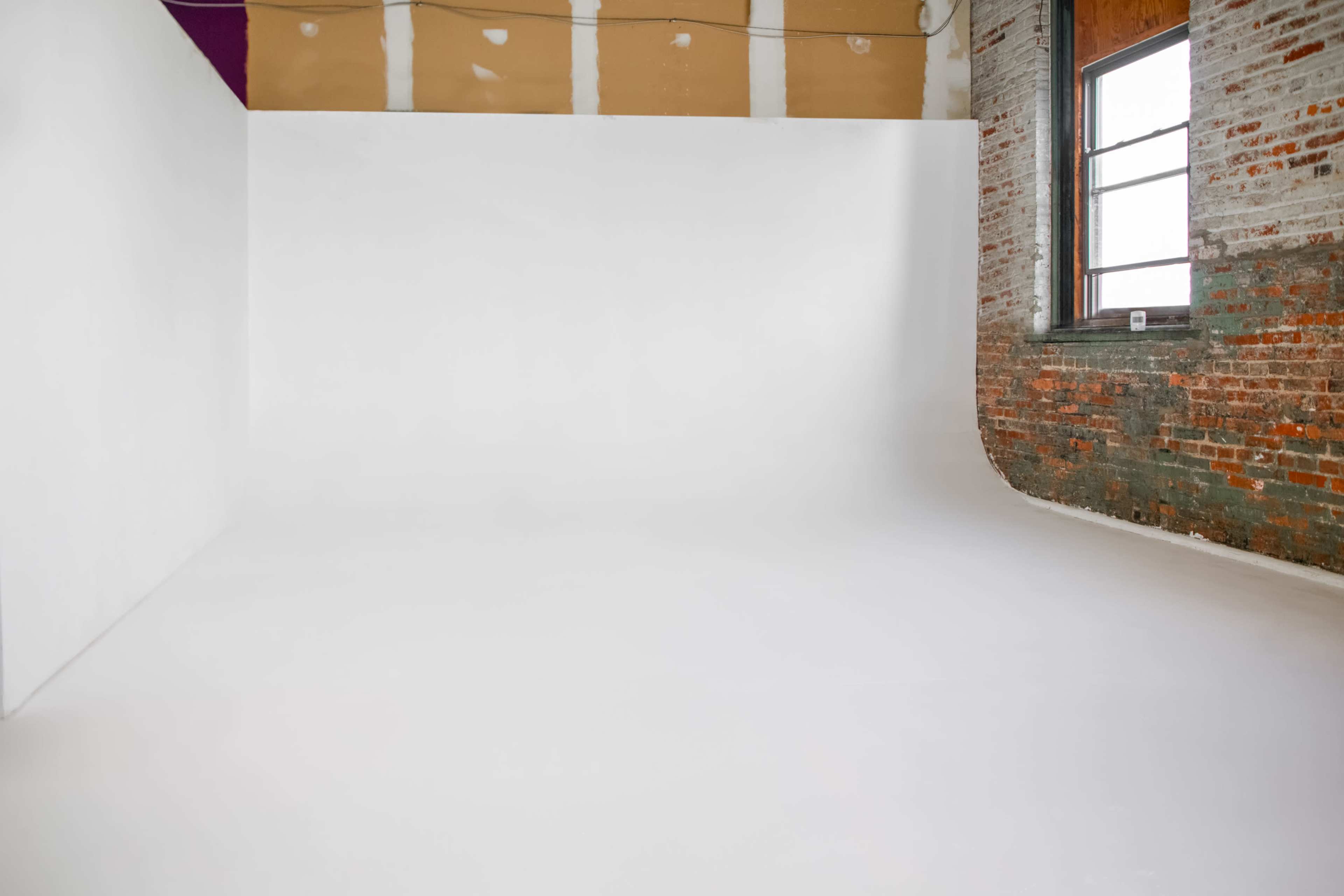 The image displays a spacious photography studio featuring a seamless white backdrop and a wall with exposed brick.