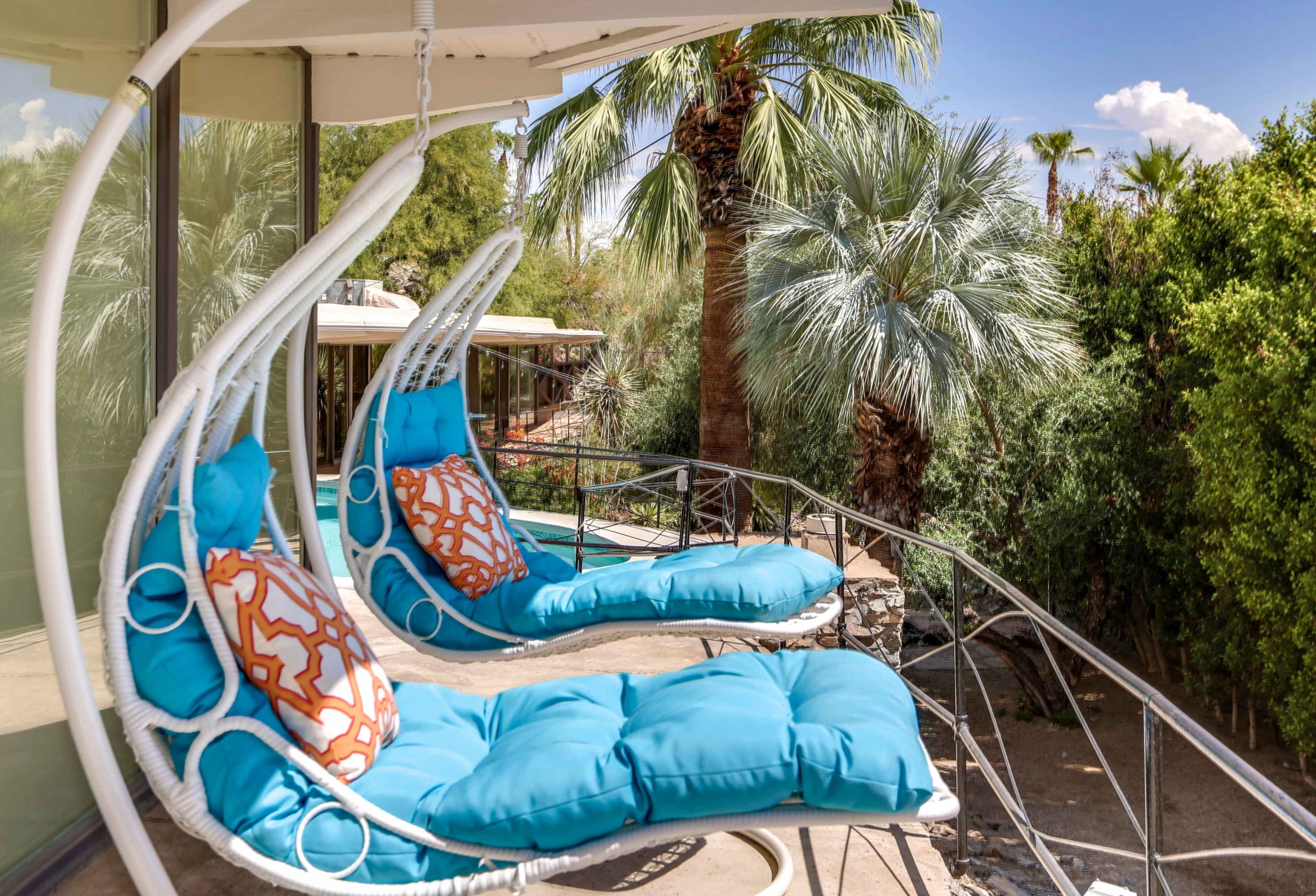 Two turquoise hanging chairs with decorative pillows are positioned on a balcony surrounded by palm trees.
