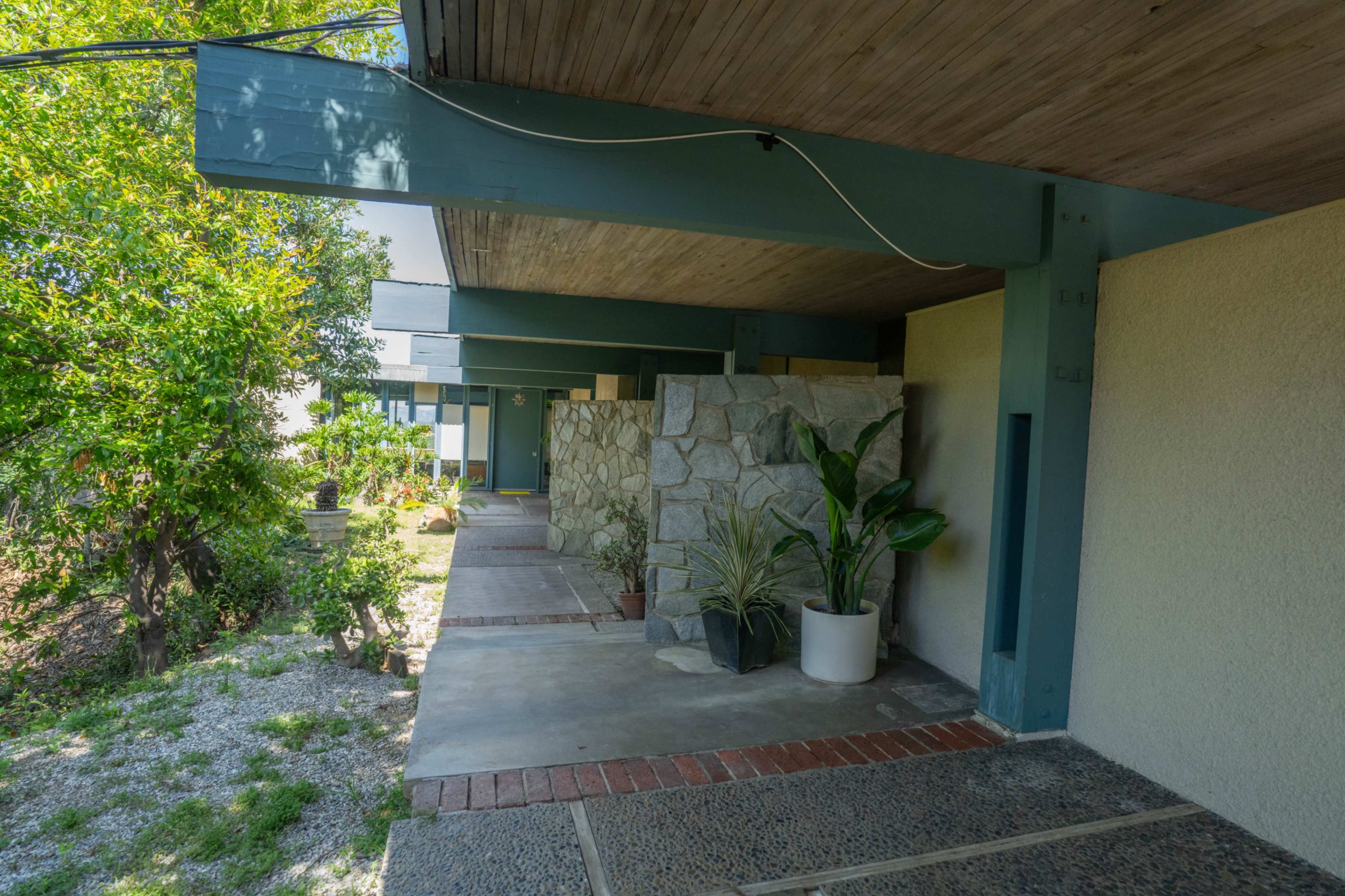 The image shows a modern building with a covered walkway, stone walls, and potted plants alongside a garden area.