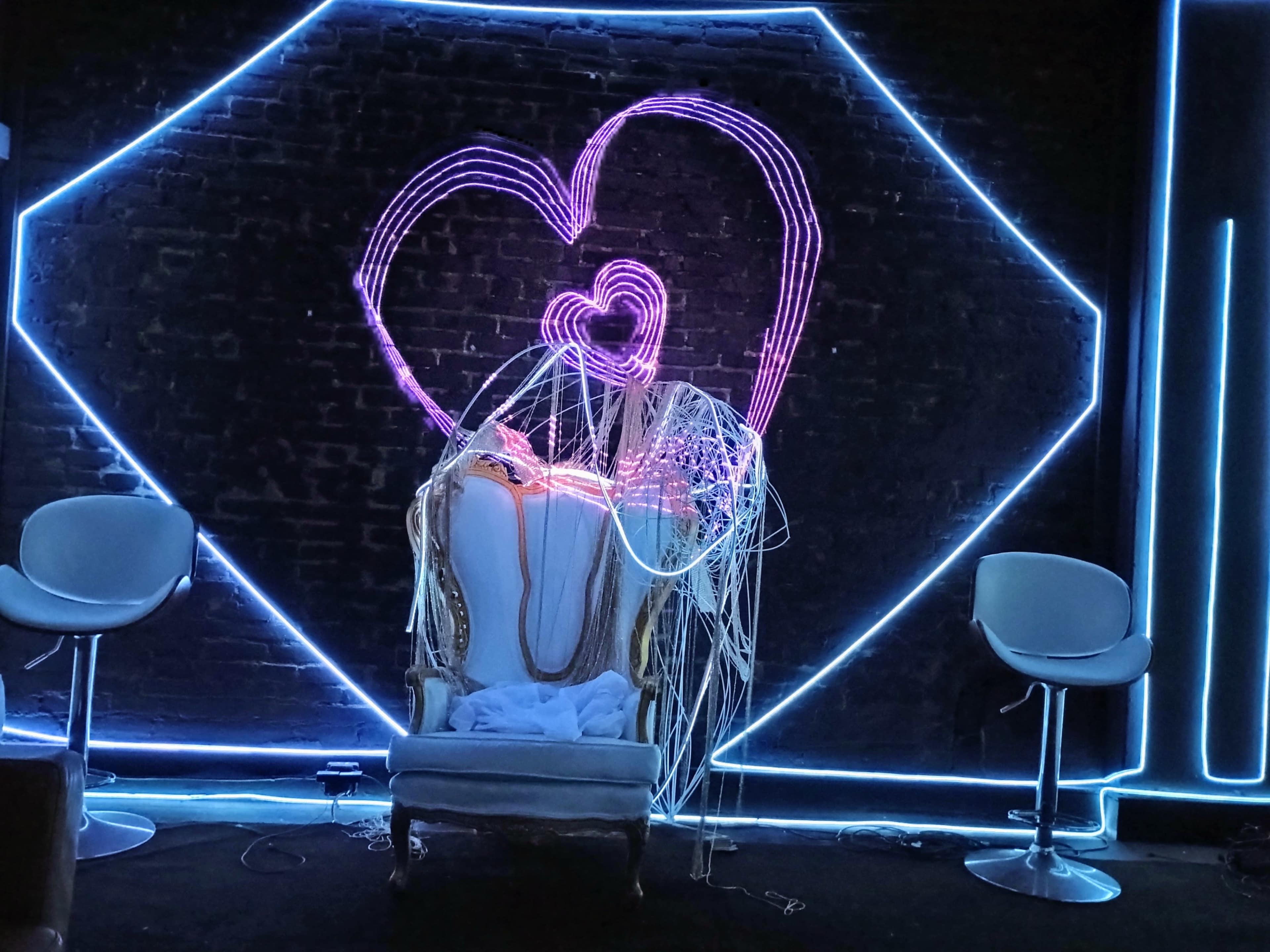 The image shows a decorative area featuring a large heart-shaped neon light installation on a brick wall, with an ornate chair in front and two modern stools nearby.