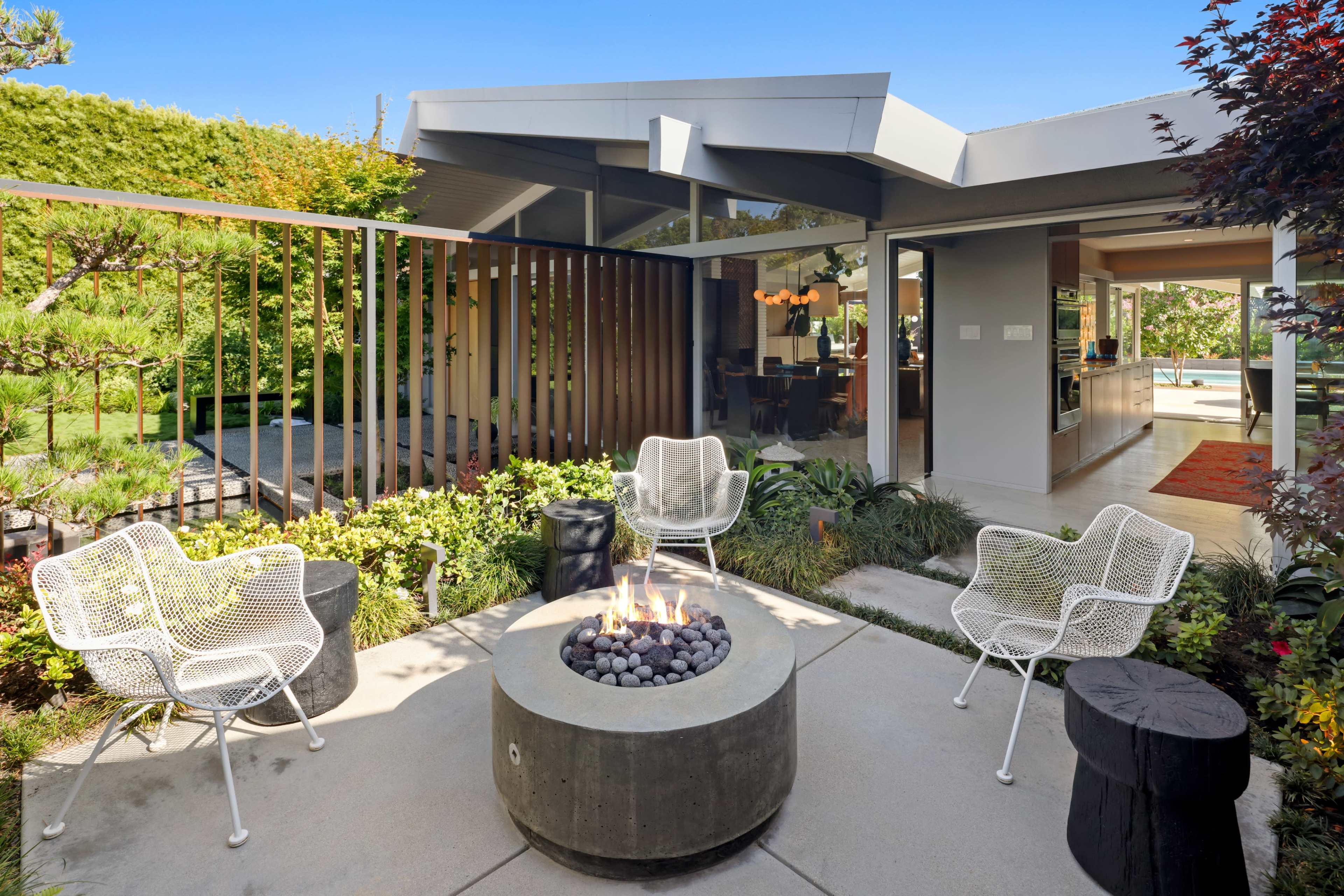 A modern outdoor seating area with four wire chairs arranged around a circular concrete fire pit, surrounded by greenery and leading to a contemporary home.