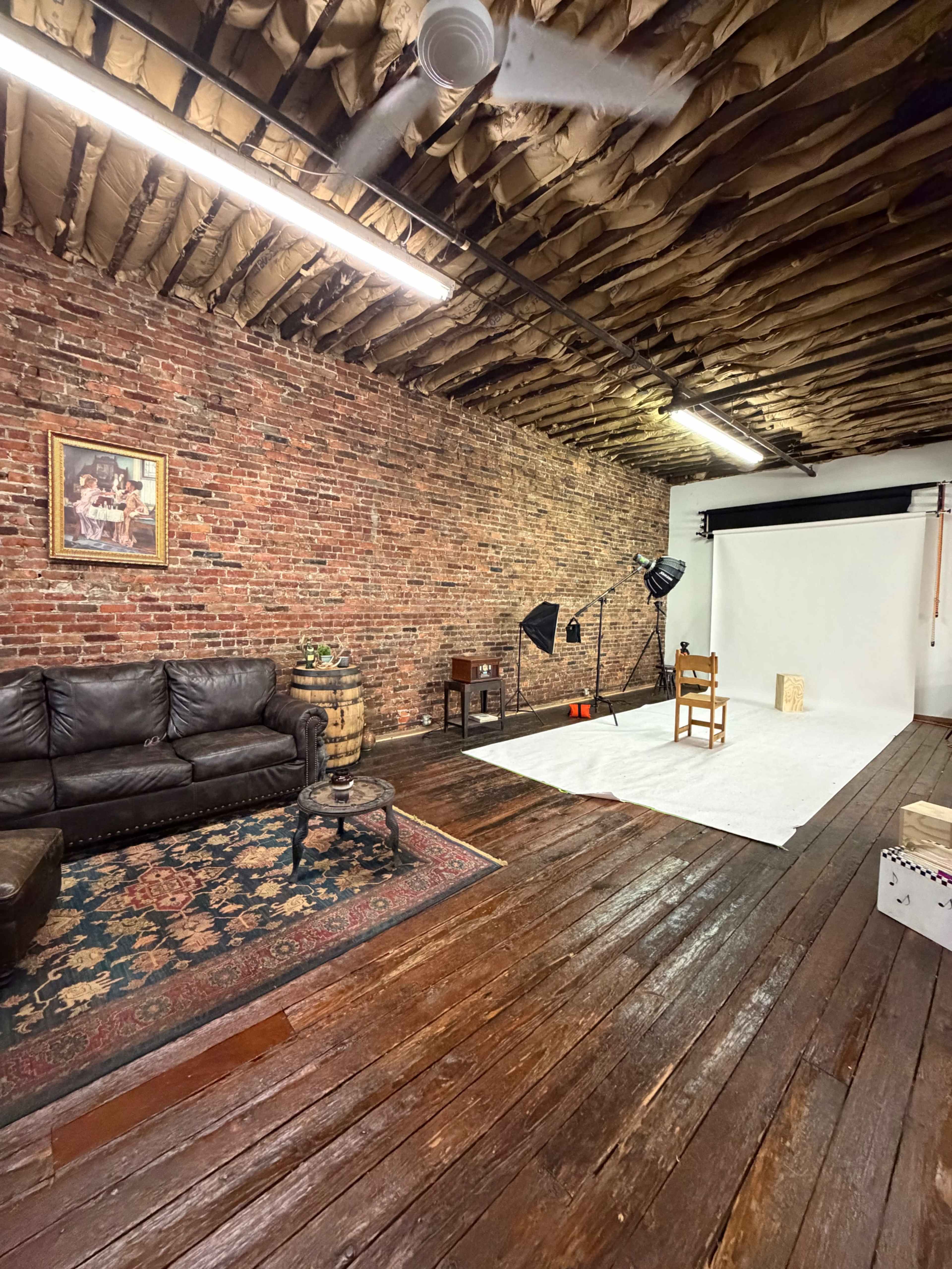 The image shows an interior space with exposed brick walls, a wooden floor, and a designated photo shoot area featuring a white backdrop, lighting equipment, and a chair.