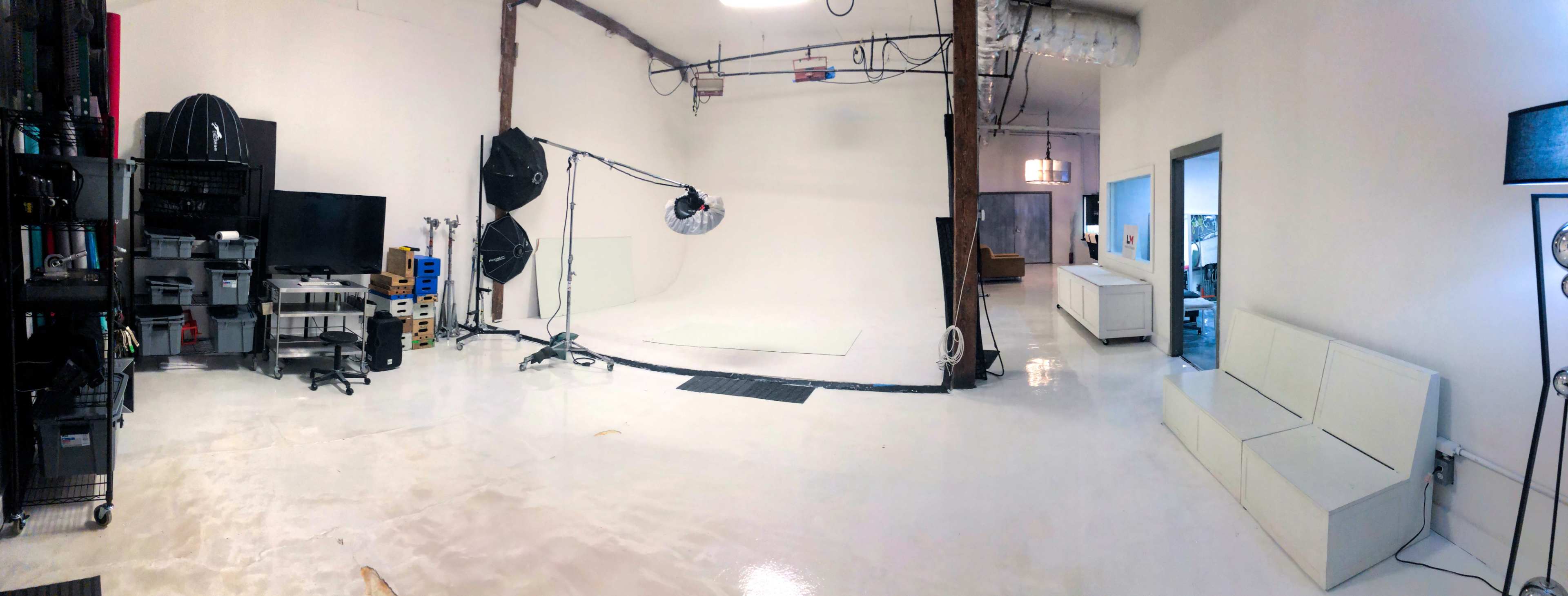 A spacious photography studio with a white backdrop, lighting equipment, a television, storage carts, and a white couch in the corner.
