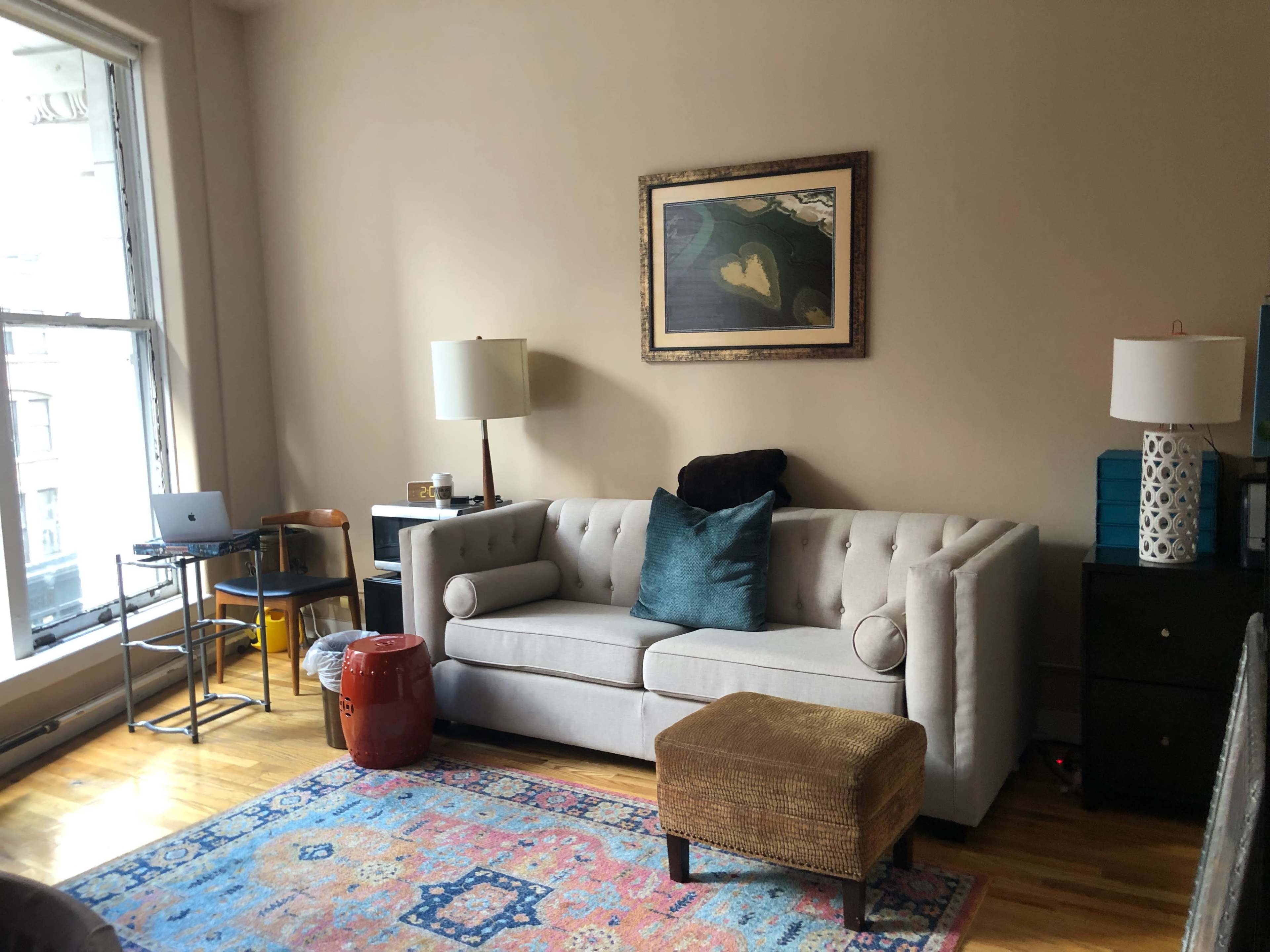 A living room with a gray tufted sofa, a small ottoman, a colorful area rug, and a desk with a laptop near a window.