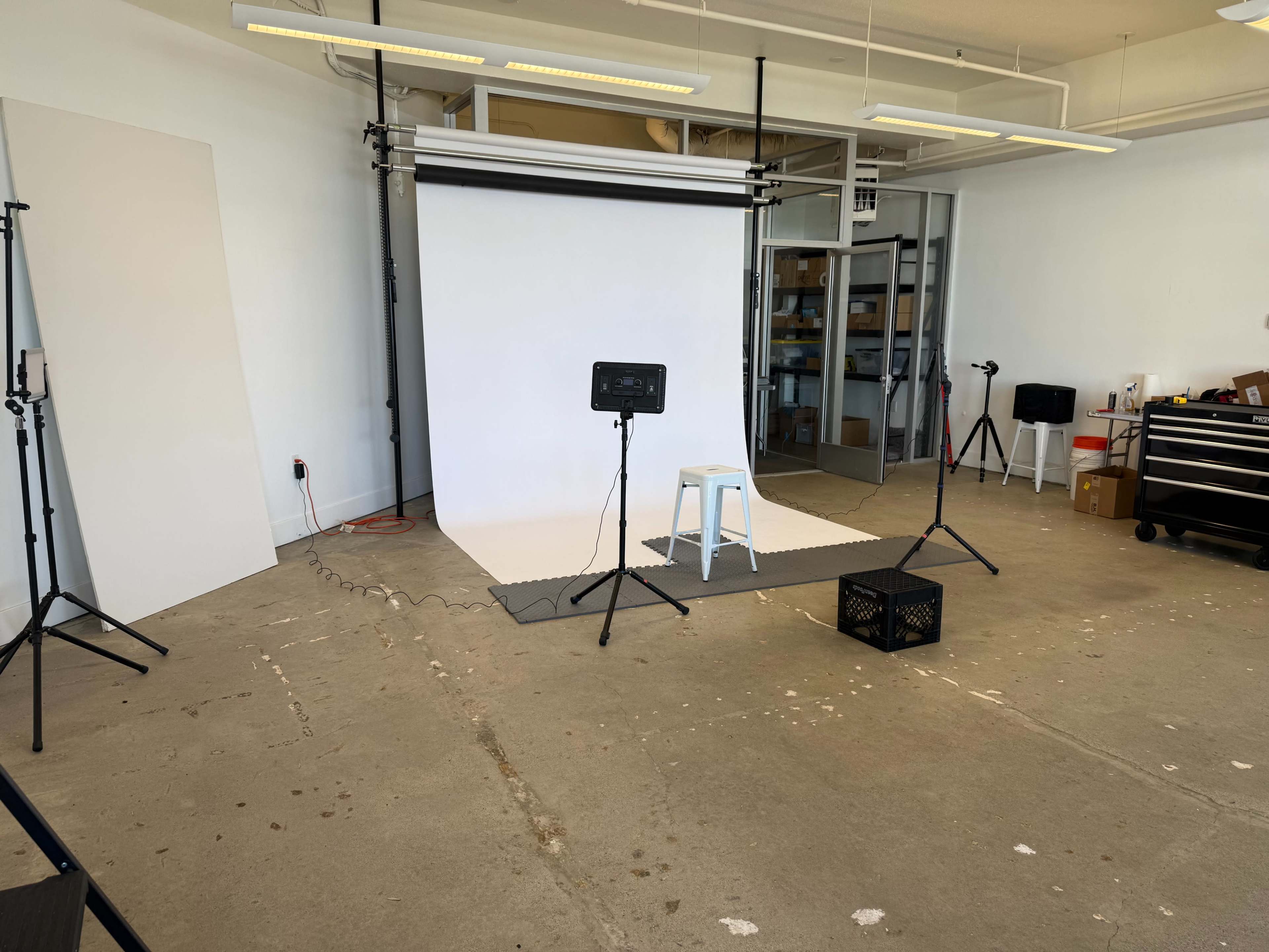 The image shows a photography studio with a white backdrop, lighting equipment, a stool, and various props arranged on a concrete floor.