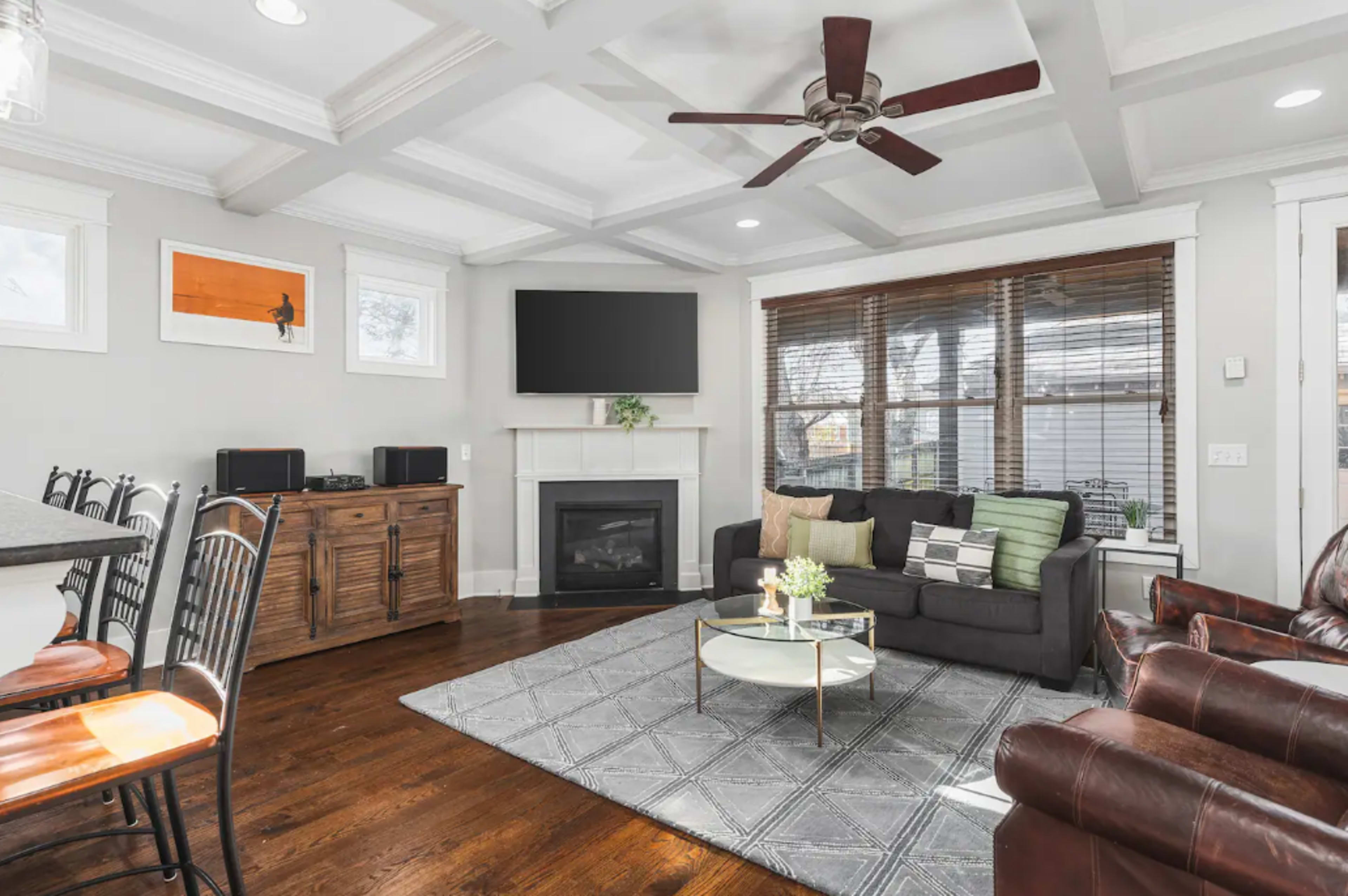 The living room features a dark sofa, a coffee table with a small plant, a flat-screen TV mounted on the wall, and a fireplace beneath a decorative shelf, with large windows allowing natural light.