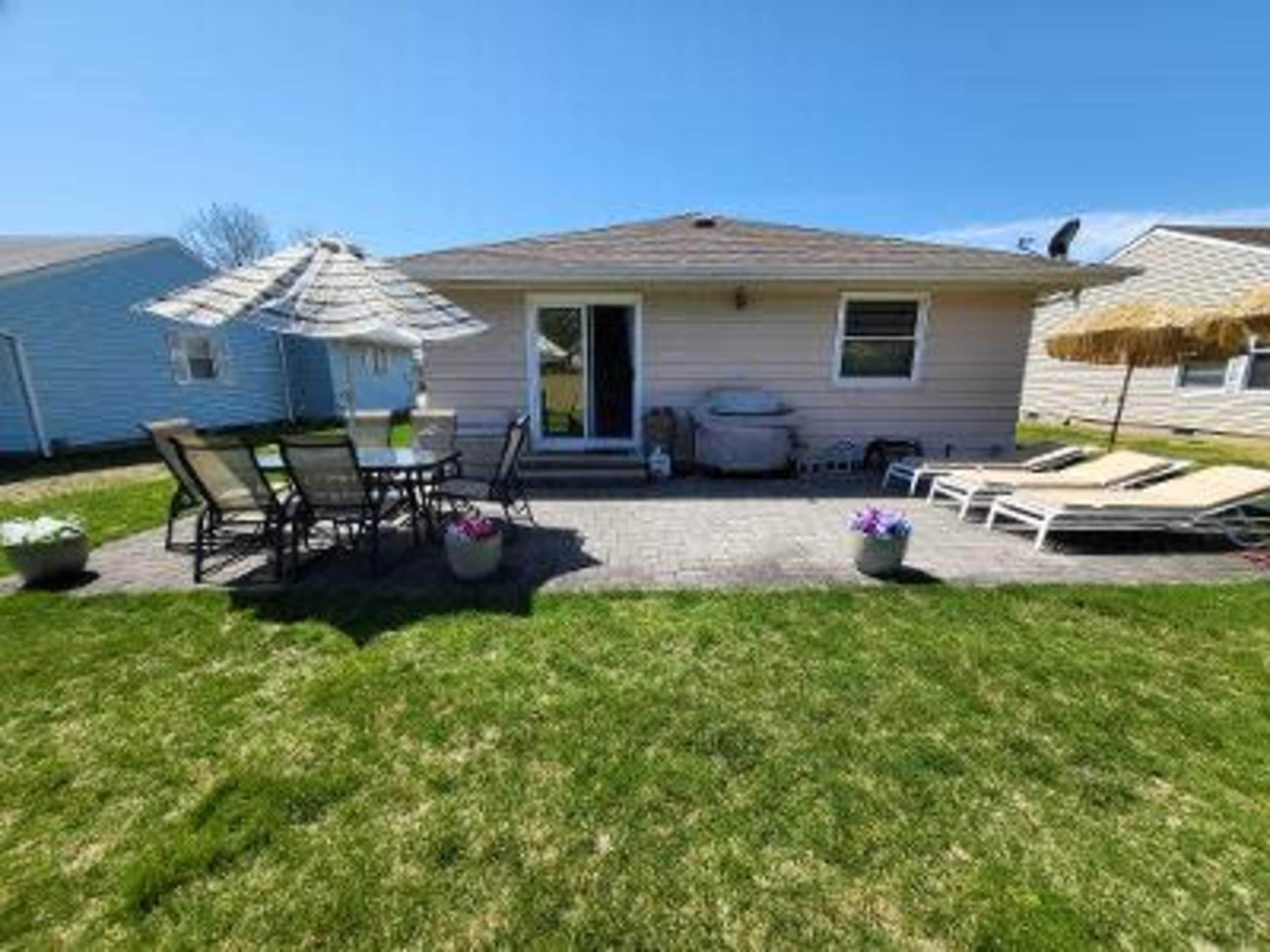 The image shows a patio area with lounge chairs, a dining set under an umbrella, and a grill, situated behind a house in a lawn setting.