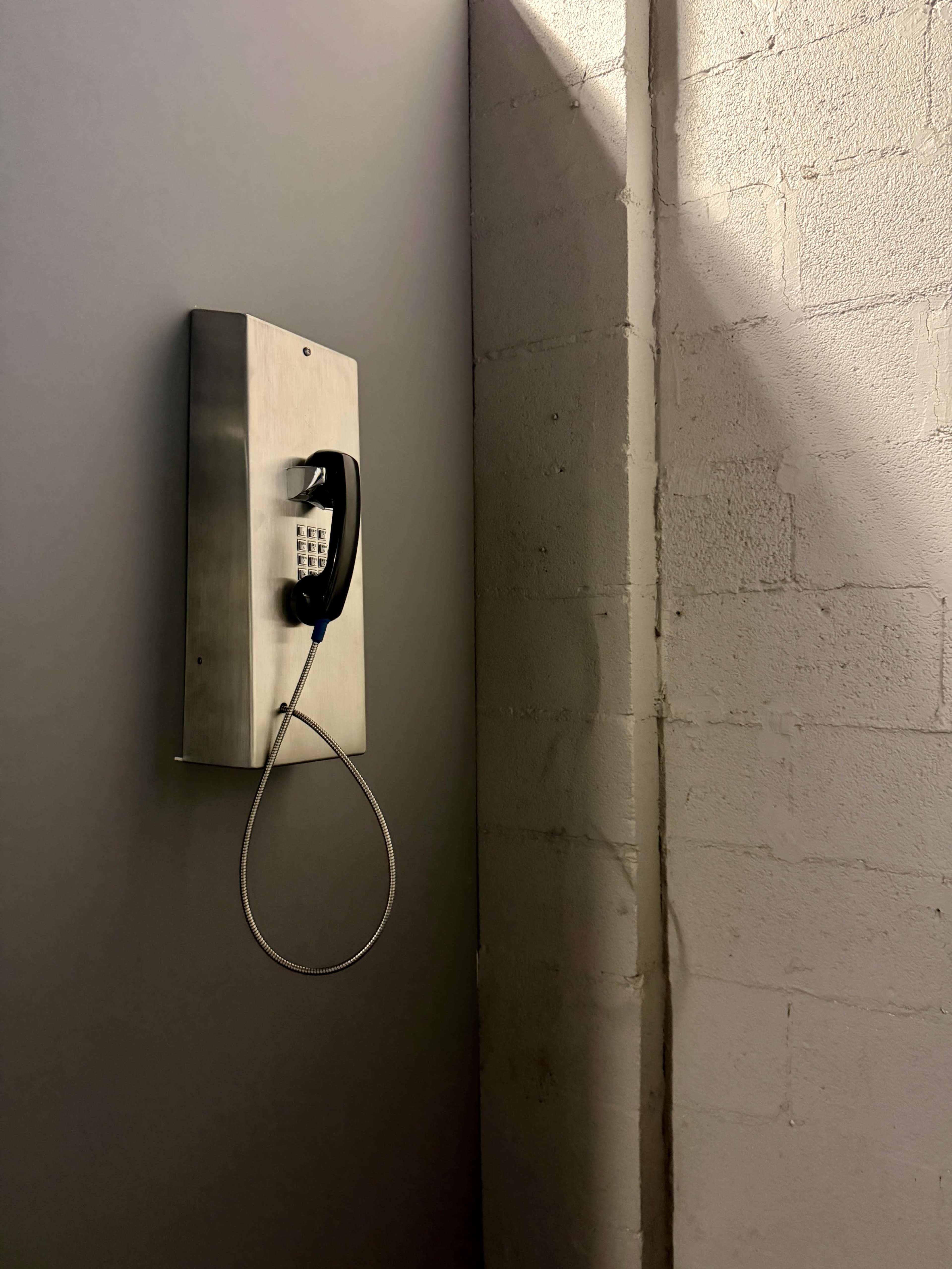 A public payphone is mounted on a gray wall next to a well-lit corner of exposed brick.
